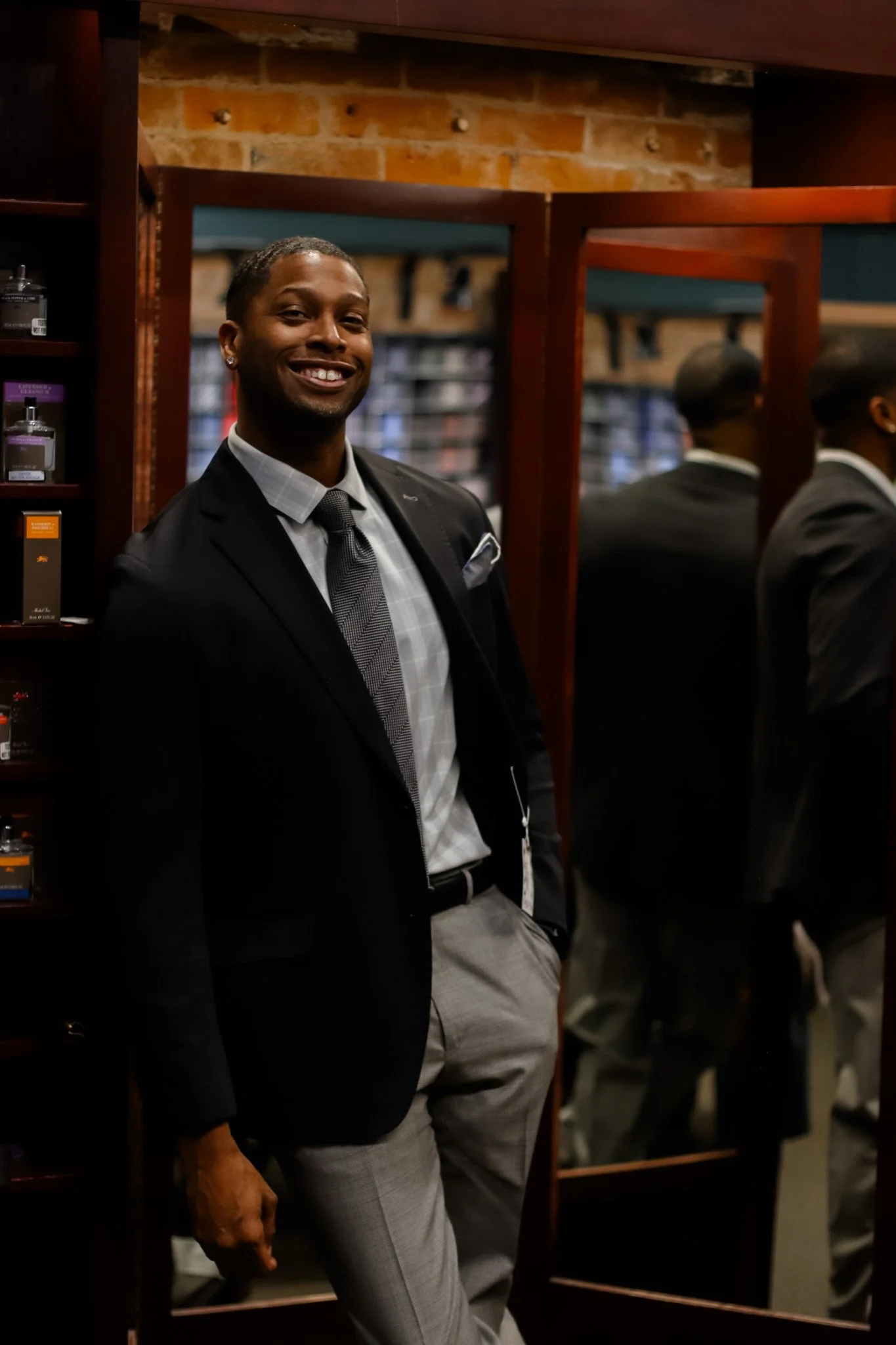 A well-dressed man in a suit and tie smiling and posing in front of a mirror inside a clothing store.
