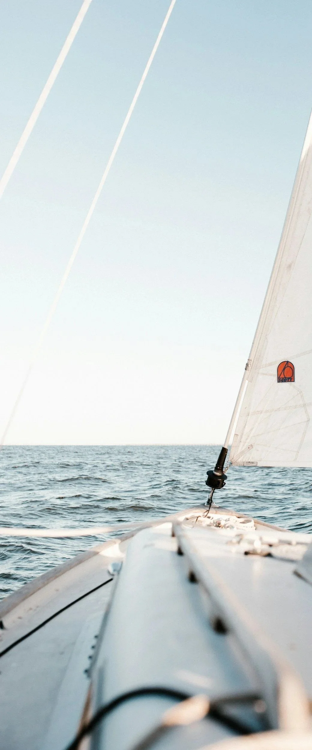 Photo from the bow of a sailing boat on open water with a white sail and calm sea.