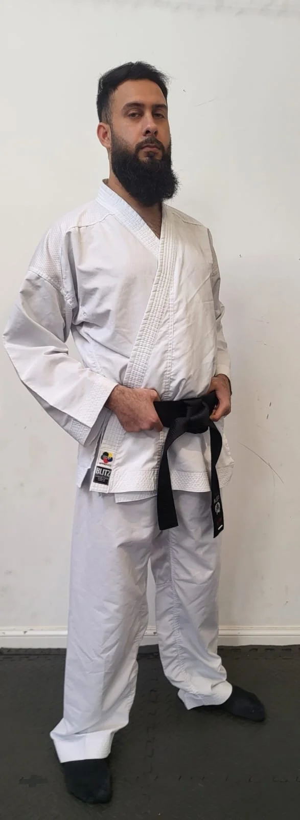 A man dressed in a white martial arts gi with a black belt standing indoors against a plain white wall.