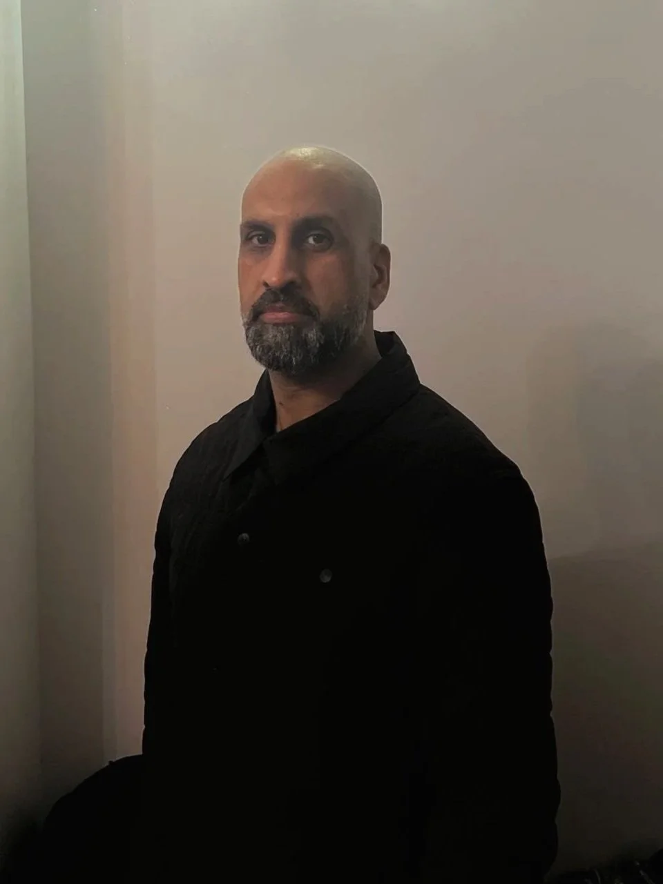A bald man with a gray beard wearing a black shirt, sitting against a plain light-colored wall.