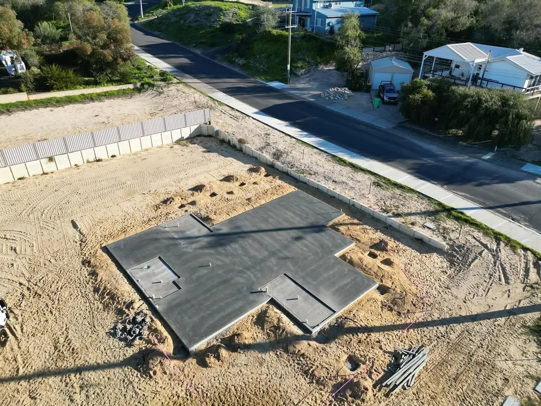 Construction site with a concrete foundation poured on the ground, surrounded by sand and dirt, with a fence and a residential neighborhood in the background.