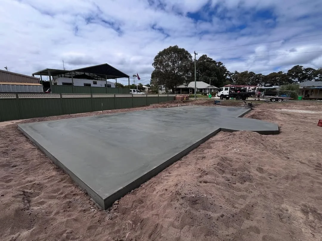 A freshly poured concrete slab on a construction site with surrounding dirt, a green fence, and residential houses in the background under a cloudy sky.