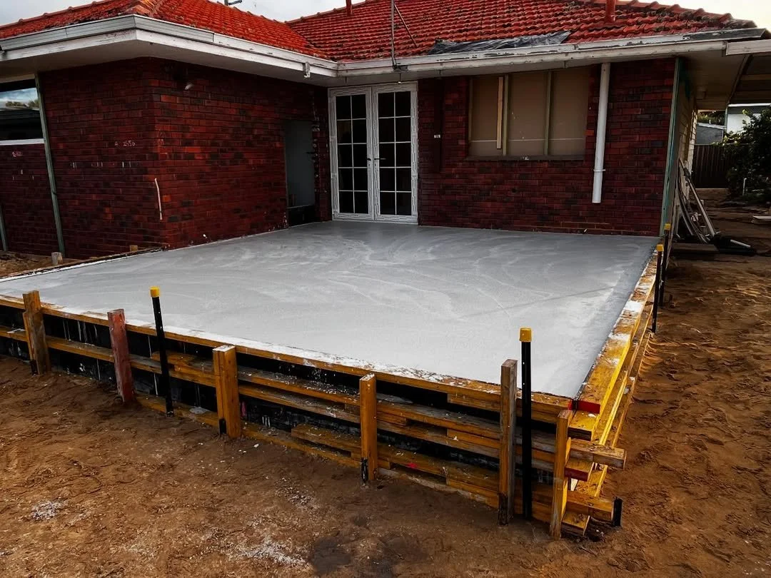 A freshly poured concrete slab in front of a red brick house, with construction framing and tools around the edges.