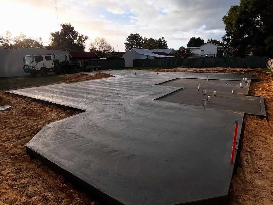 Construction site with freshly poured concrete foundation on a residential lot, with pipes protruding from the concrete for plumbing or utilities, and a truck and houses in the background at sunset.