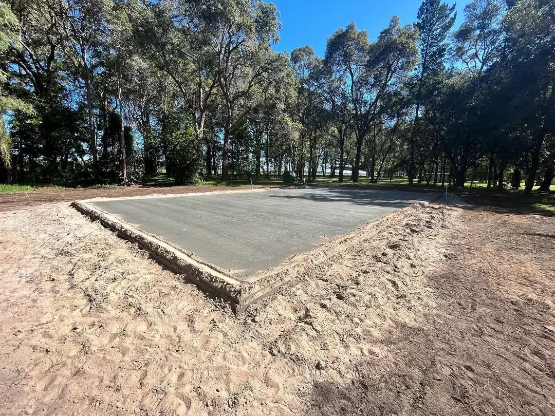 Freshly poured concrete slab on a construction site surrounded by dirt with trees and blue sky in the background.