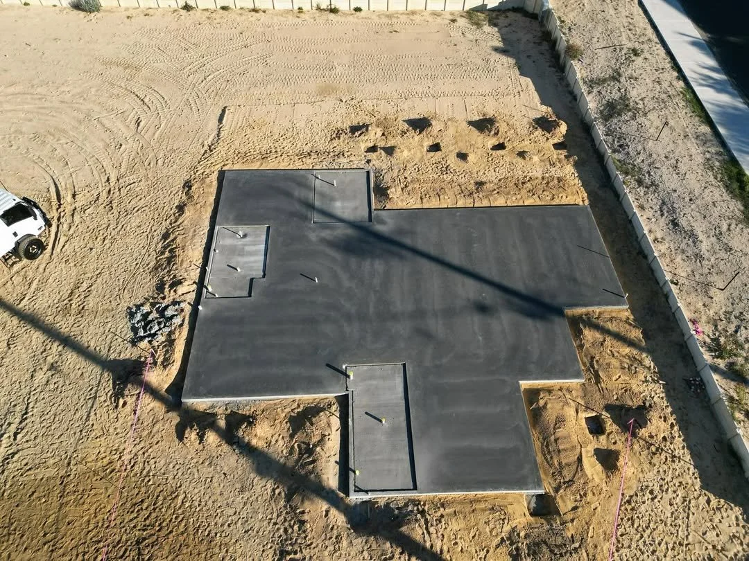 A construction site showing a newly poured concrete foundation in the shape of a house, with piles of gravel and dirt surrounding it. There are some construction markings and a white vehicle parked nearby.