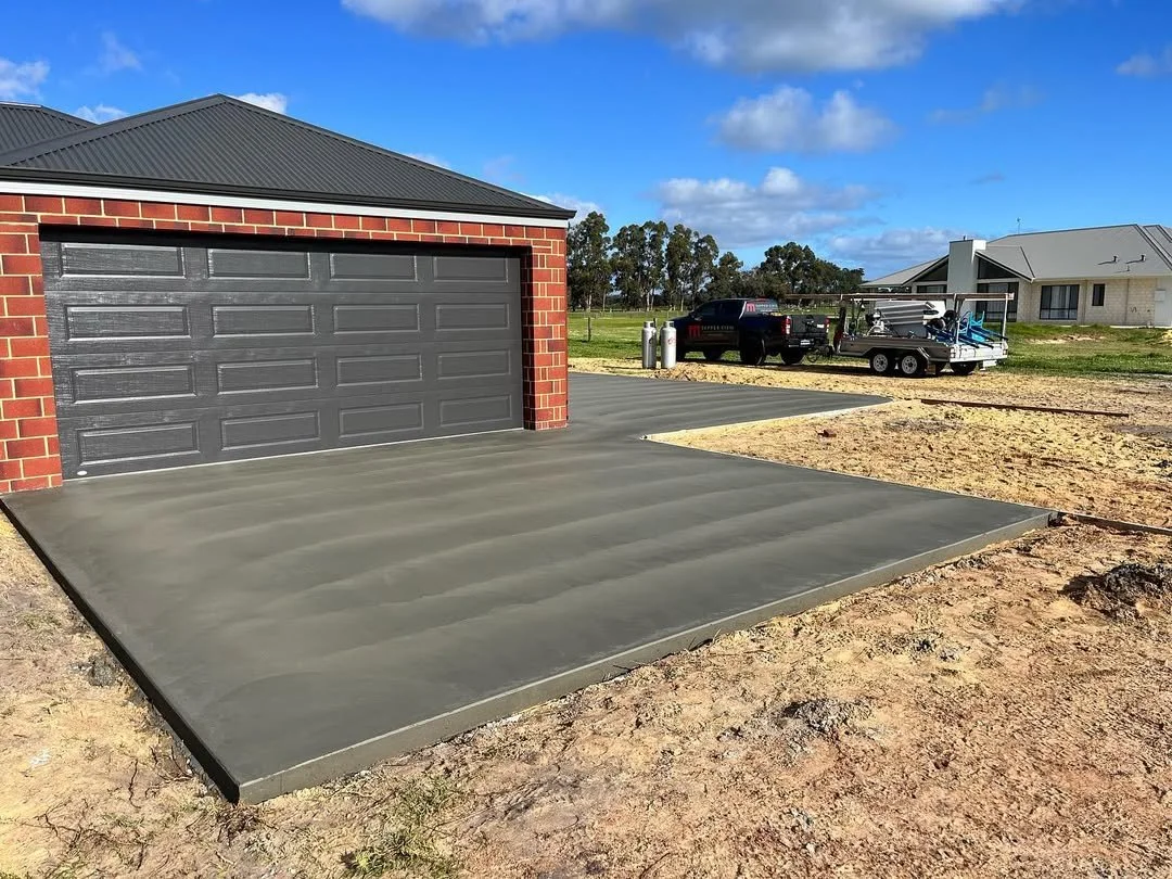 Freshly poured concrete driveway in front of a brick garage with a closed gray garage door, surrounded by dirt and grass, with a pickup truck and trailer nearby.
