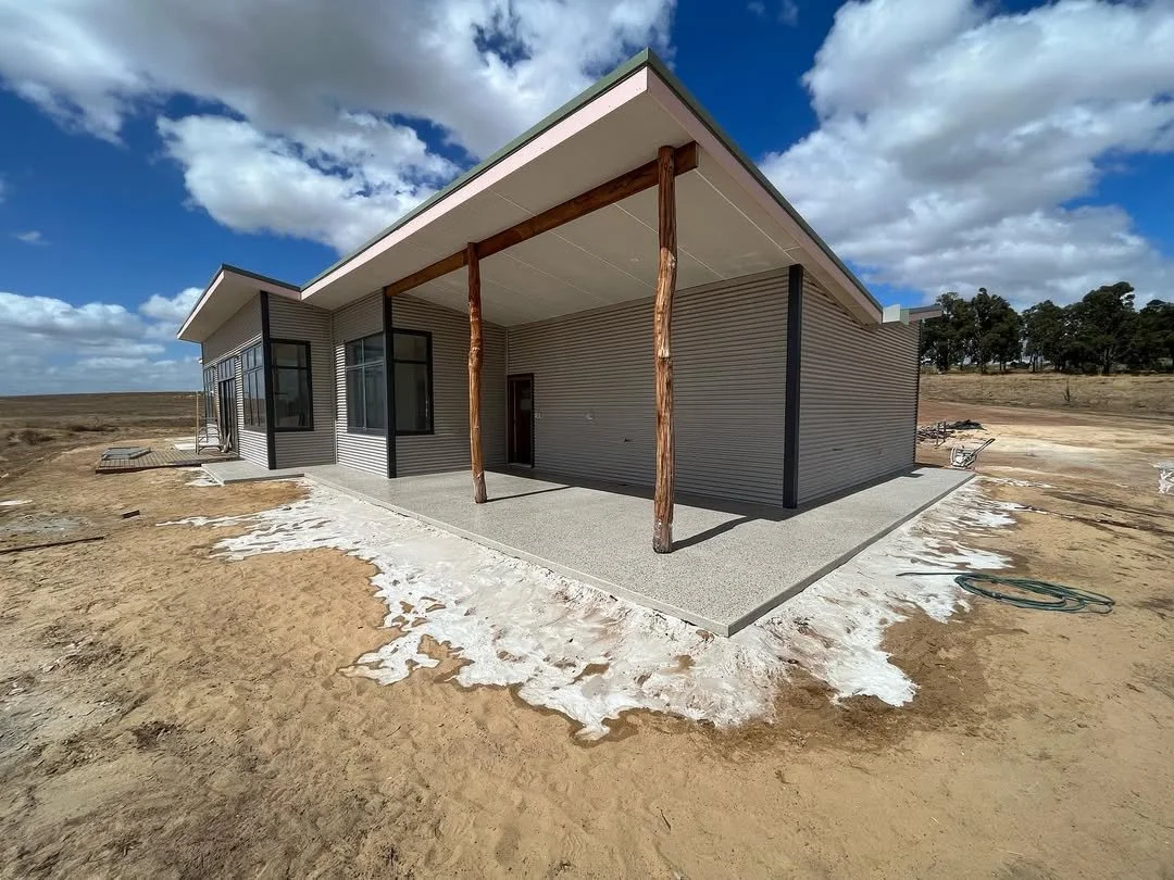 Newly constructed modern house with vinyl siding and large windows, featuring a covered porch with wooden poles, situated on a dirt lot under a partly cloudy sky.
