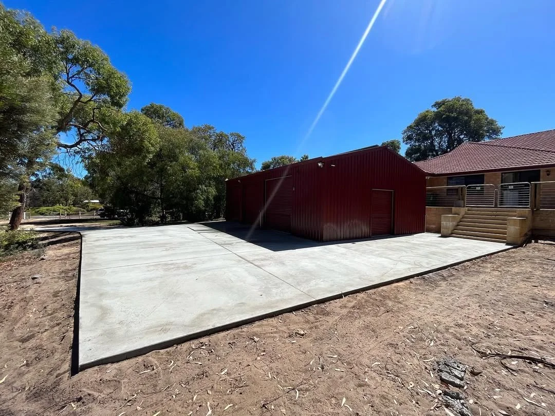 Concrete patio in front of a red shed with a roller door, next to a house with a tiled roof and a small set of stairs, with trees in the background and a clear blue sky.
