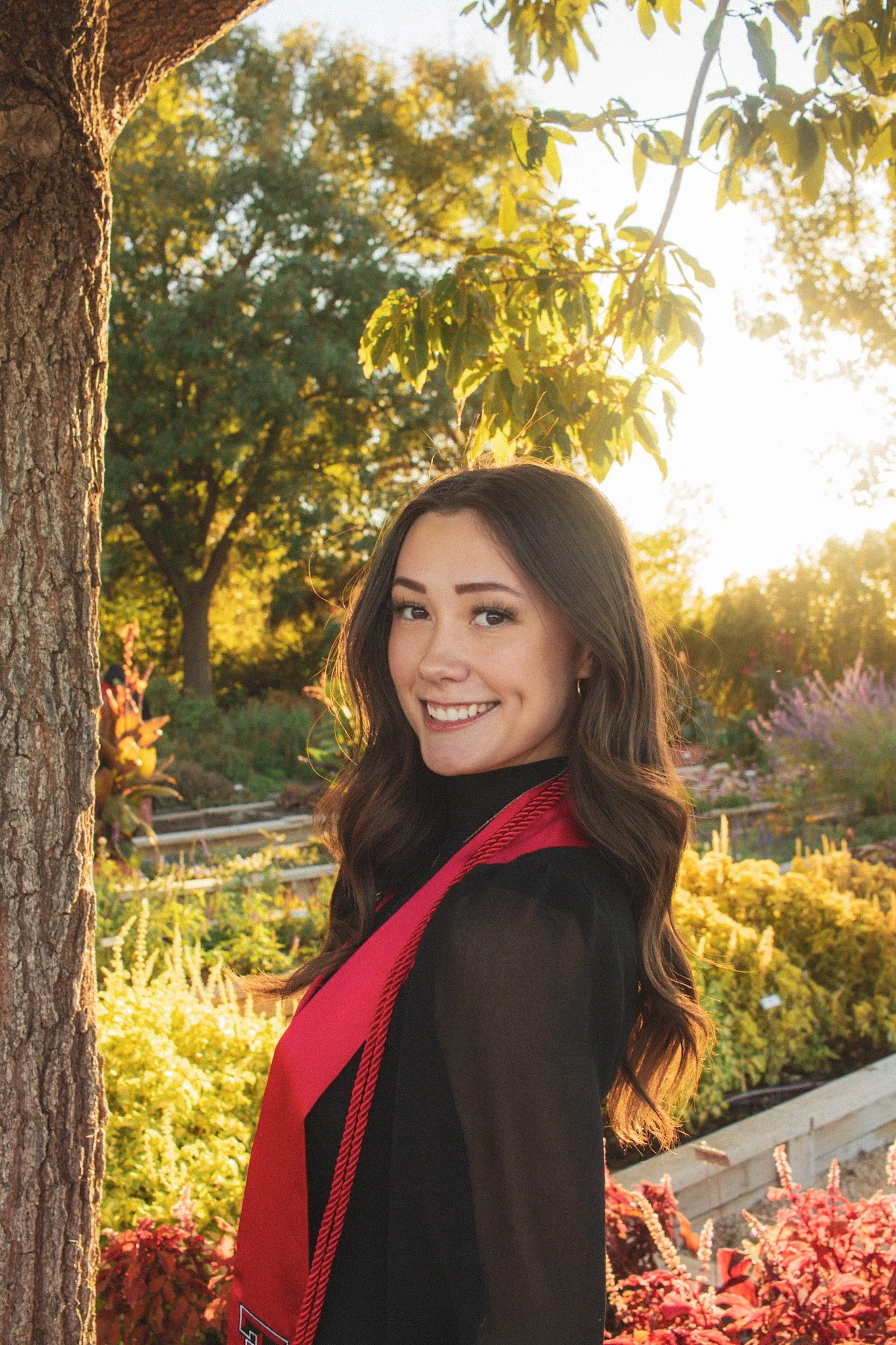 Young woman in a graduation gown smiling outdoors in a garden, with sunlight and trees in the background.