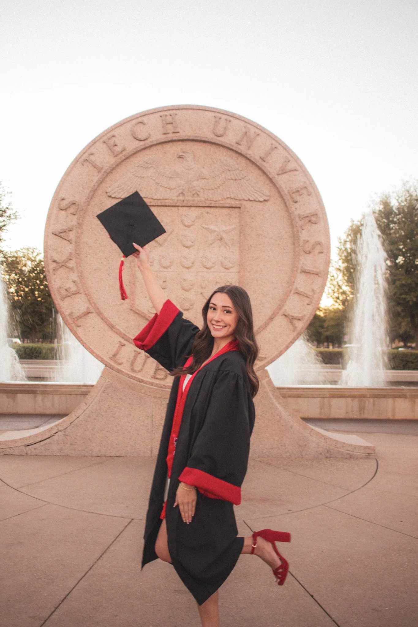 A young woman in a graduation gown and red heels holding a diploma cover, celebrating in front of a large Texas State University monument during sunset.