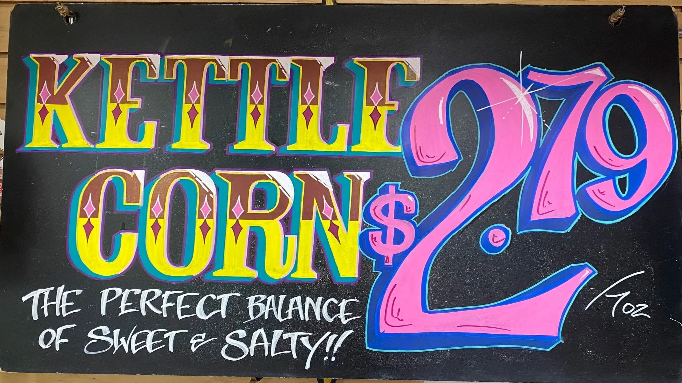 Colorful end-cap sign promoting kettle corn with a large pink and blue 3D-style number 2 and dollar sign. The sign says 'KETTLE CORN 2$' with the phrase 'The perfect balance of sweet & salty!!' written below in white cursive text.