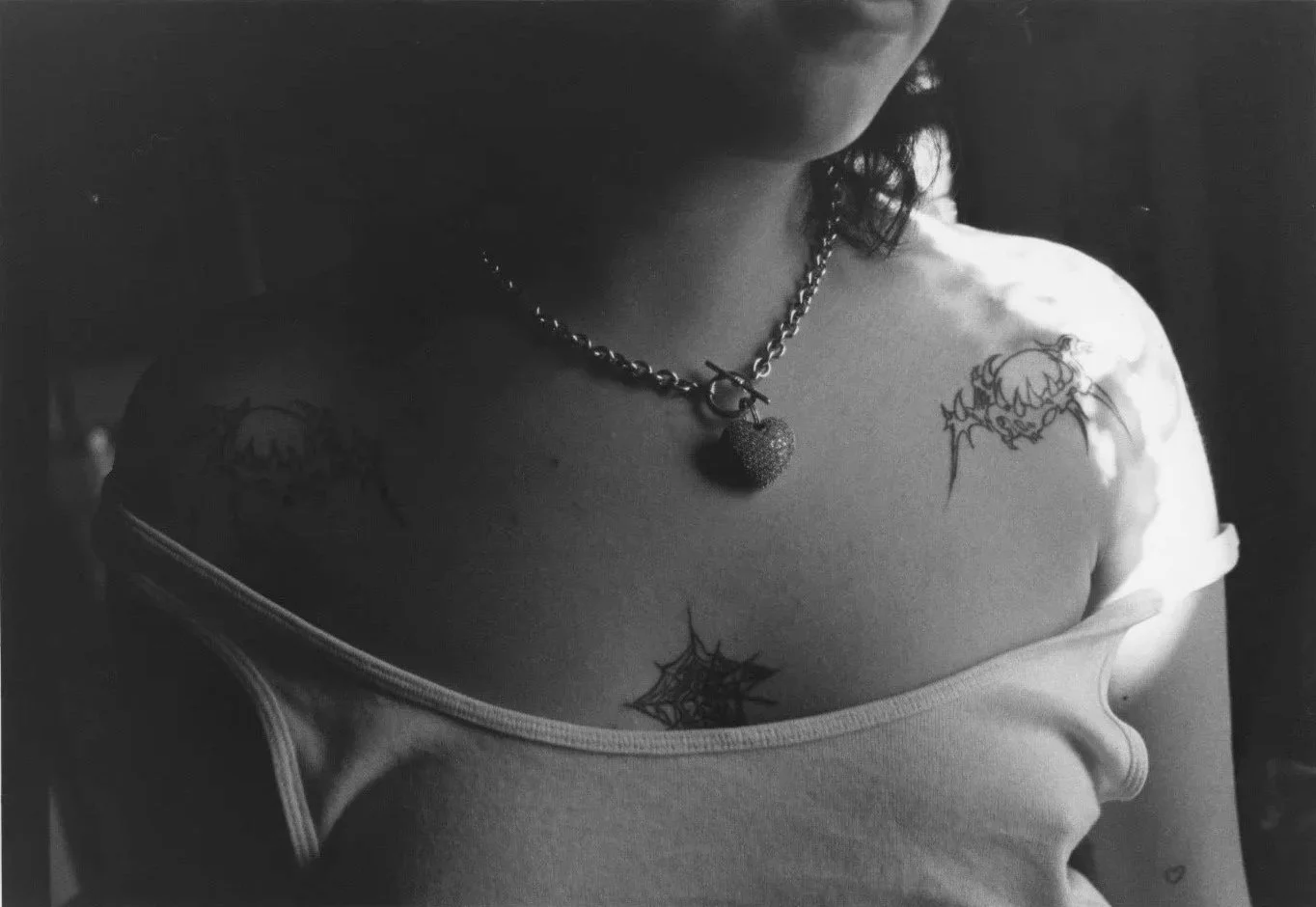 Close-up black and white photo of a person's upper chest, showing tattoos of spiderwebs and a creature, and a necklace with a strawberry pendant, with part of a tank top visible.