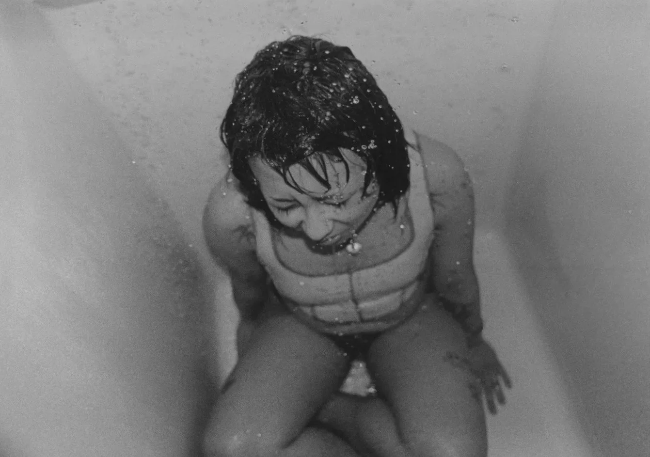 A woman with short dark hair, glasses, and a striped shirt sitting in a bathtub, captured from above in a black-and-white photo.