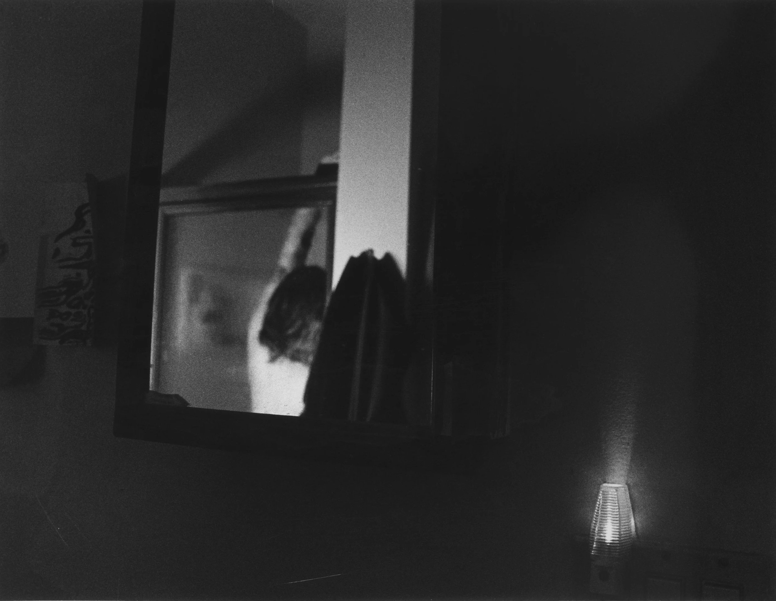A black and white photo of a room with a square mirror mounted on the wall, reflecting a person hanging clothes. To the right of the mirror, there is a small lamp emitting soft light.