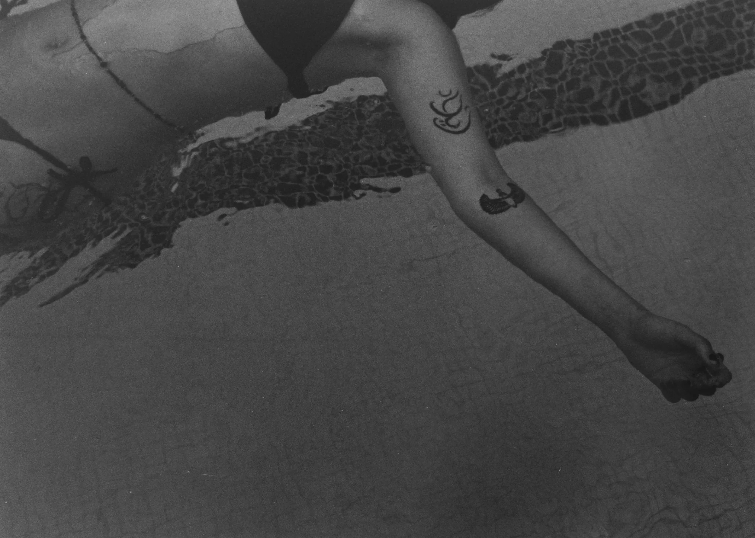 A person in a swimsuit taking a mirror selfie in a dimly lit room, with tattoos on their arm and leg, and a leopard print towel hanging on a wall.