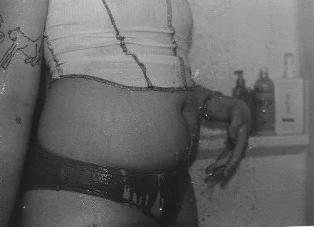 Close-up of a person's midsection wearing a cropped top and low-rise underwear, with a cluttered background including bottles and containers.