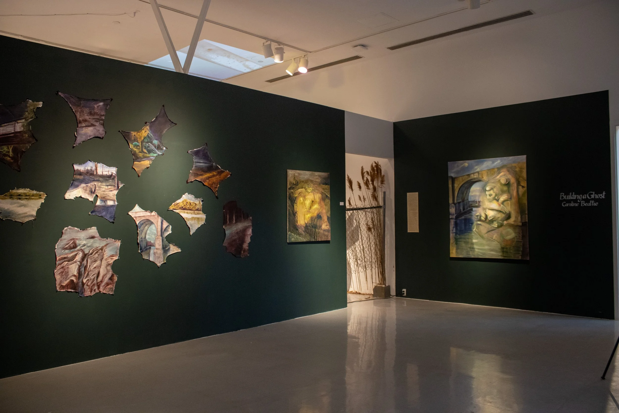 An art gallery exhibit featuring painted artworks and hanging fabric or paper pieces on dark green walls, with light coming from a skylight.