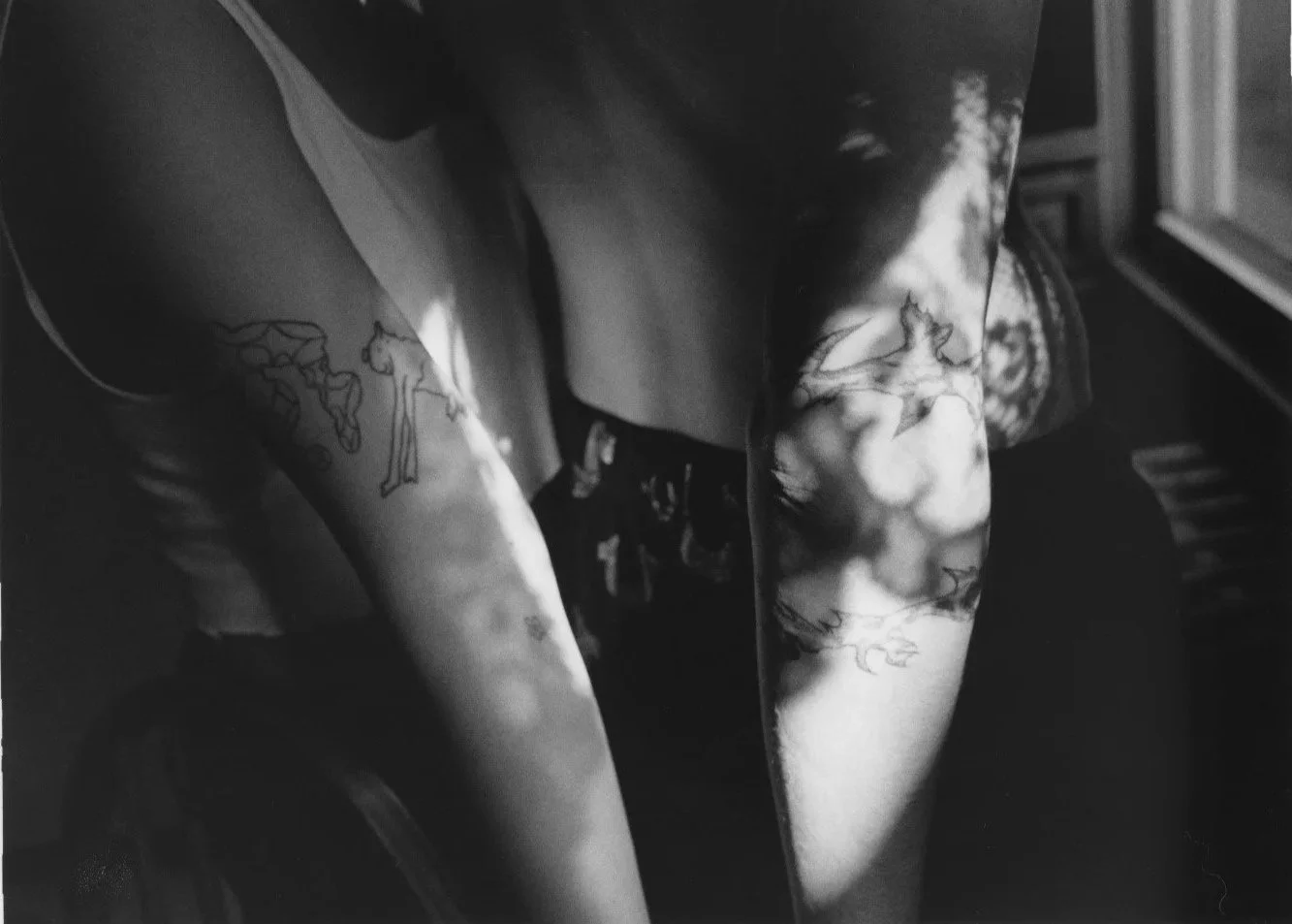 Black and white photograph of a person with tattoos on their arms and torso, with abstract and animal designs, standing near a window with sunlight casting shadows.