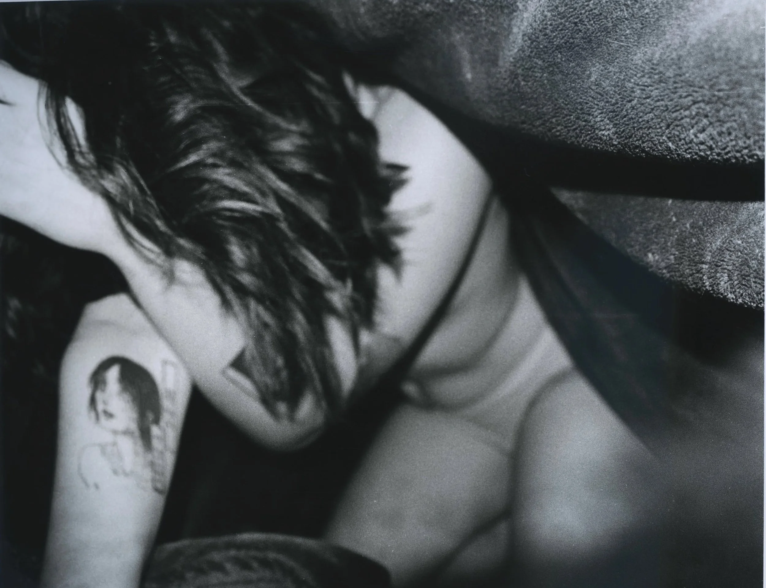 A woman with wavy hair is leaning on her arm on a surface, with her head turned down, showing a tattoo on her upper arm.