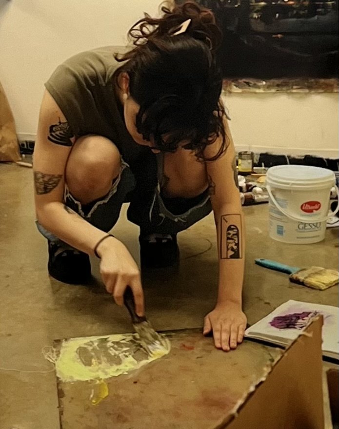 A woman with tattoos is kneeling on the floor, working on a craft project with a small spatula or palette knife. She has black curly hair with a white hair clip and is wearing an olive green t-shirt and ripped black jeans. Various supplies and a large white bucket are around her, suggesting an art or craft activity.