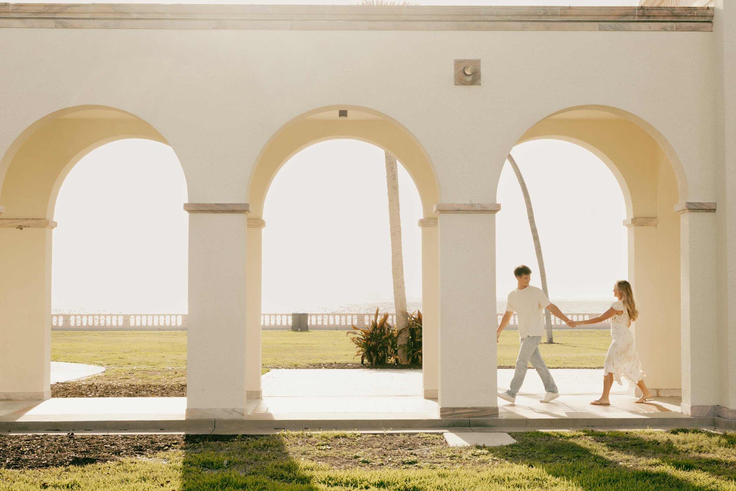 A young man and a young girl holding hands and walking in front of arches overlooking the bay of Sarasota, FL, with the sun shining brightly as he leads her.
