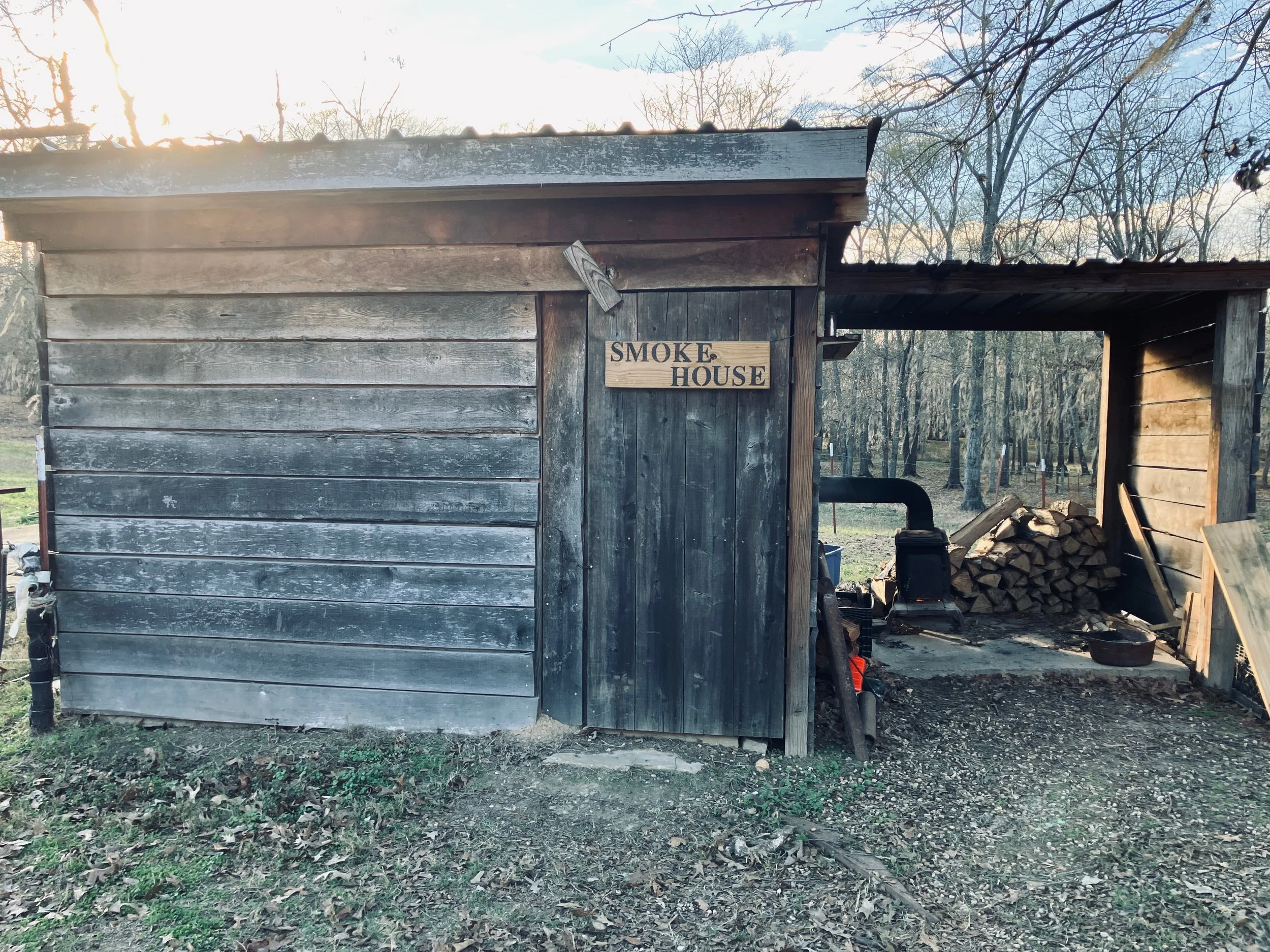 An outdoor wooden smokehouse with a sign that reads 'Smoke House', stored firewood piled inside, and a wood-burning stove pipe. The setting is a wooded area with trees in the background.