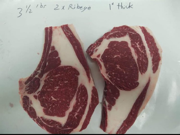 Two pieces of ribeye steak with marbling on a white surface, with handwritten notes reading "3 1/2" bbr 2 x Ribeye 1" Thick".