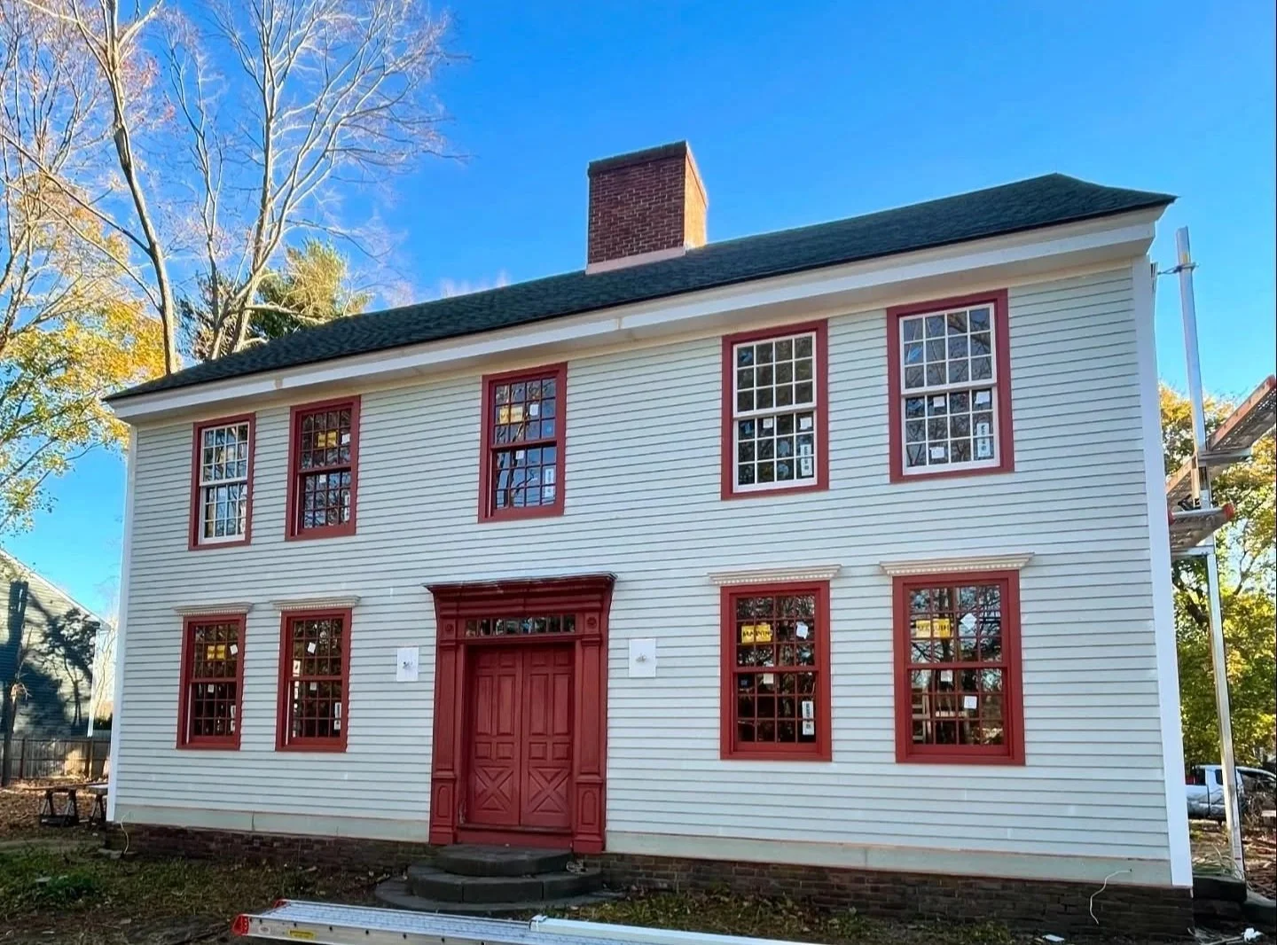 A two-story house with white siding and red trim around the windows and door, a green roof, and a brick chimney, surrounded by trees with autumn colors and a clear blue sky. Heartwood Renovations. West Hartford, CT
