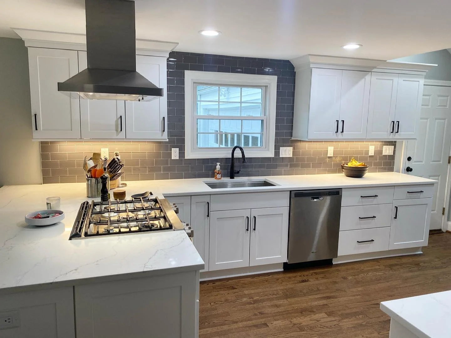 Modern kitchen with white cabinets, gray subway tile backsplash, stainless steel dishwasher, and a window above the sink. There is a stove with a stainless steel range hood, and marble countertops. Items on the counter include utensils in a holder, a bowl of bananas, and a dish with candy. 