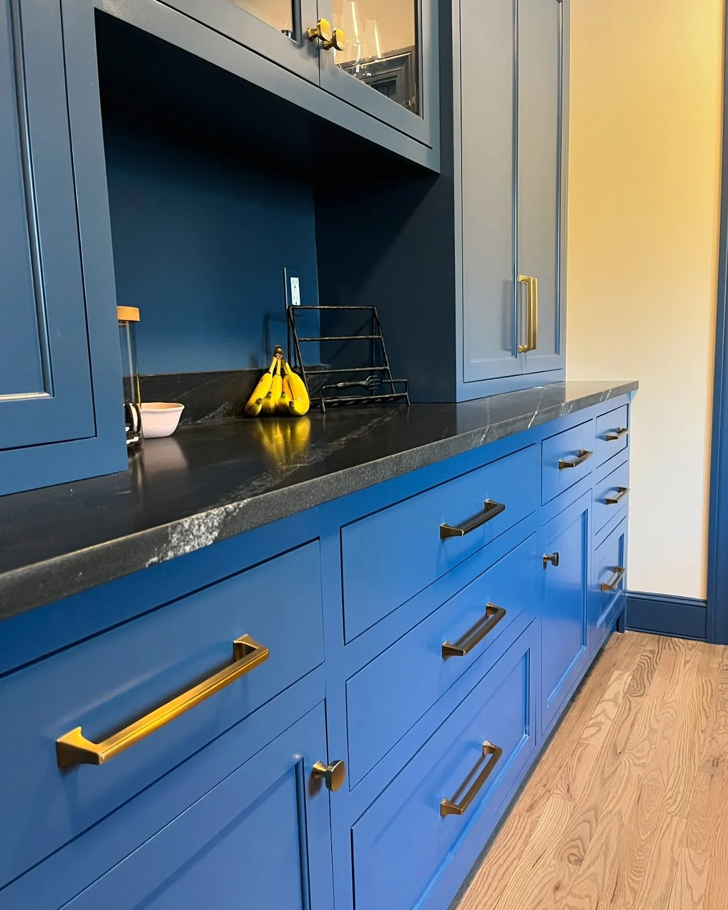 Blue kitchen cabinets with black countertop, gold handles, a banana bunch, a black wire rack, and a wood floor. Heartwood Renovations. West Hartford, CT