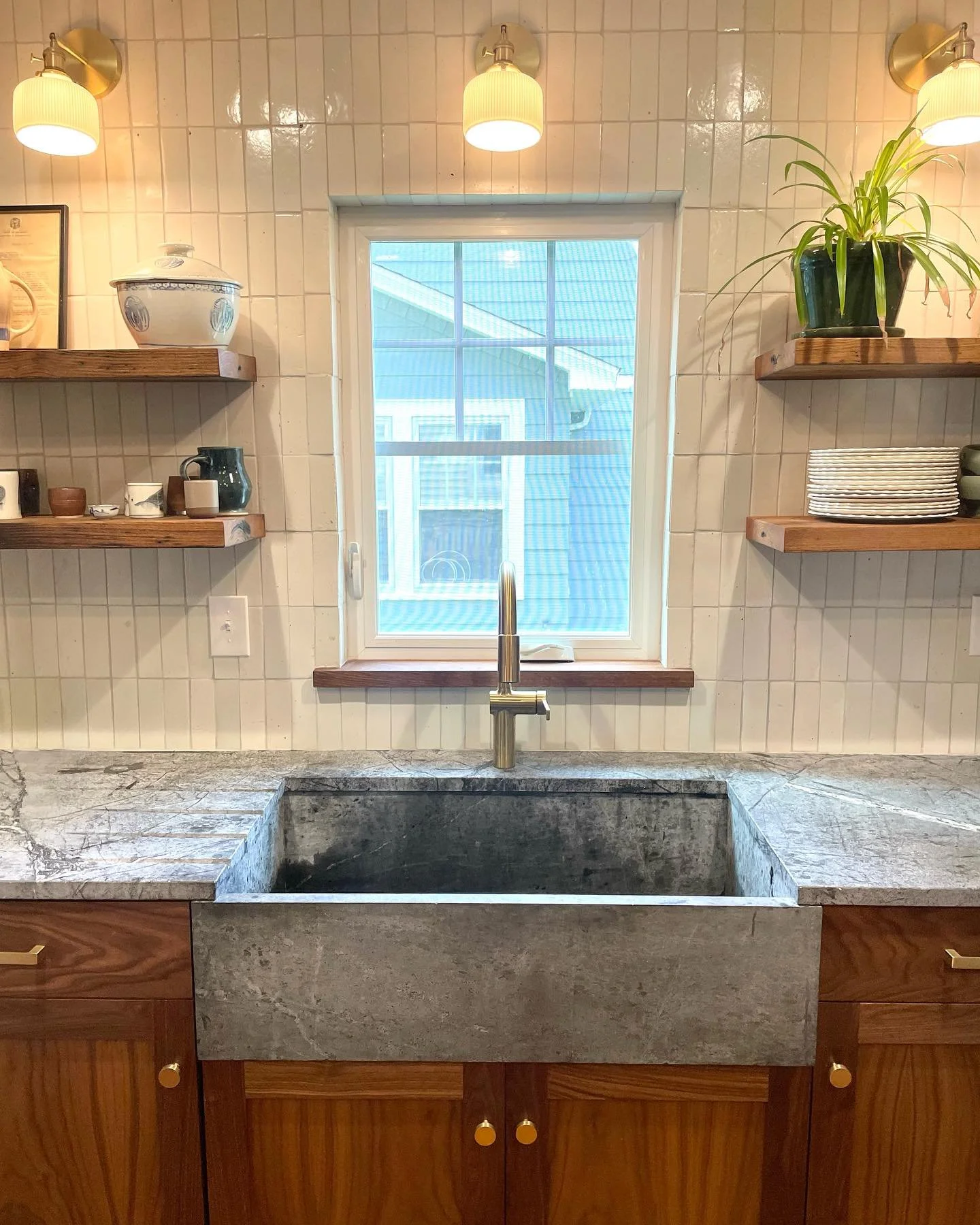 A rustic kitchen sink with a stone countertop, brass faucet, and wooden cabinets beneath. Two wooden shelves hold dishes, cups, a potted plant, and decorative jars, with a window above the sink allowing natural light into the space. Heartwood Renovat