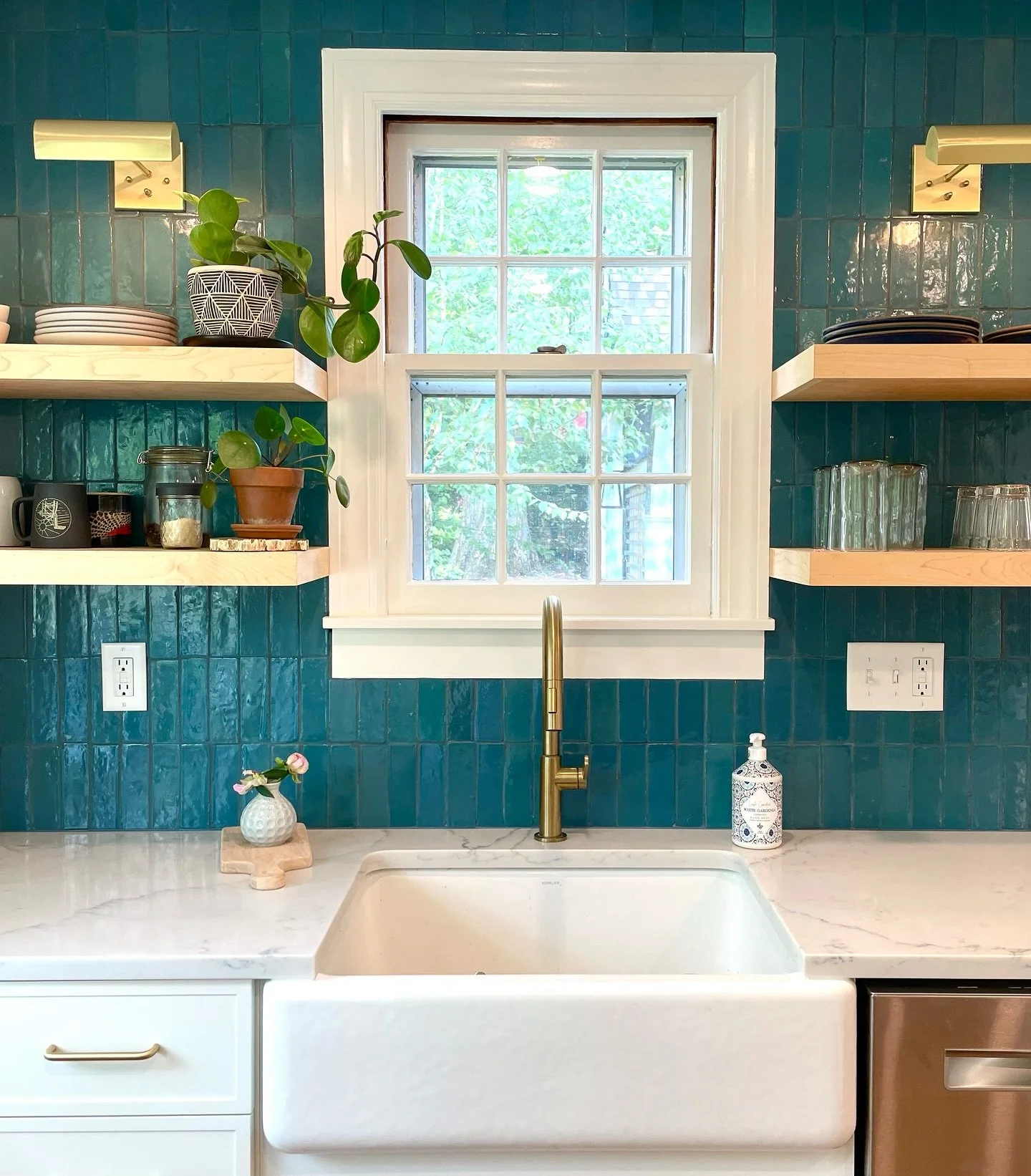 Kitchen with teal tile walls, white farmhouse sink, gold faucet, marble countertop, open wooden shelves with dishes, glassware, and plants, window with white trim, soap dispenser, and small vase with flowers. Heartwood Renovations. West Hartford, CT