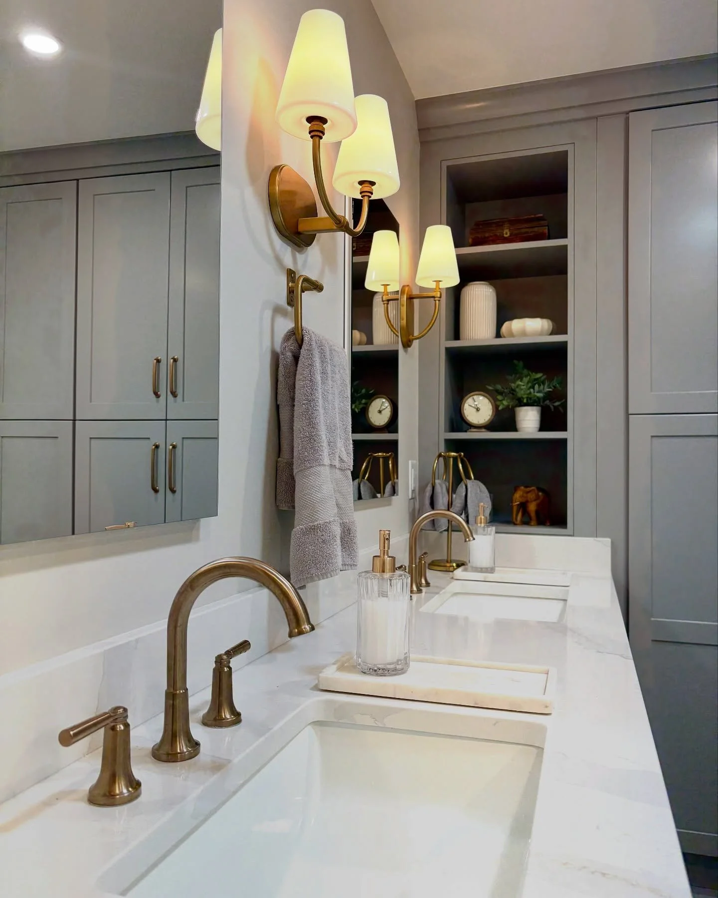 Bathroom with a marble countertop, two sinks, gold fixtures, wall-mounted gold lamps, gray cabinets, open shelves with decor and supplies. Heartwood Renovations. West Hartford, CT