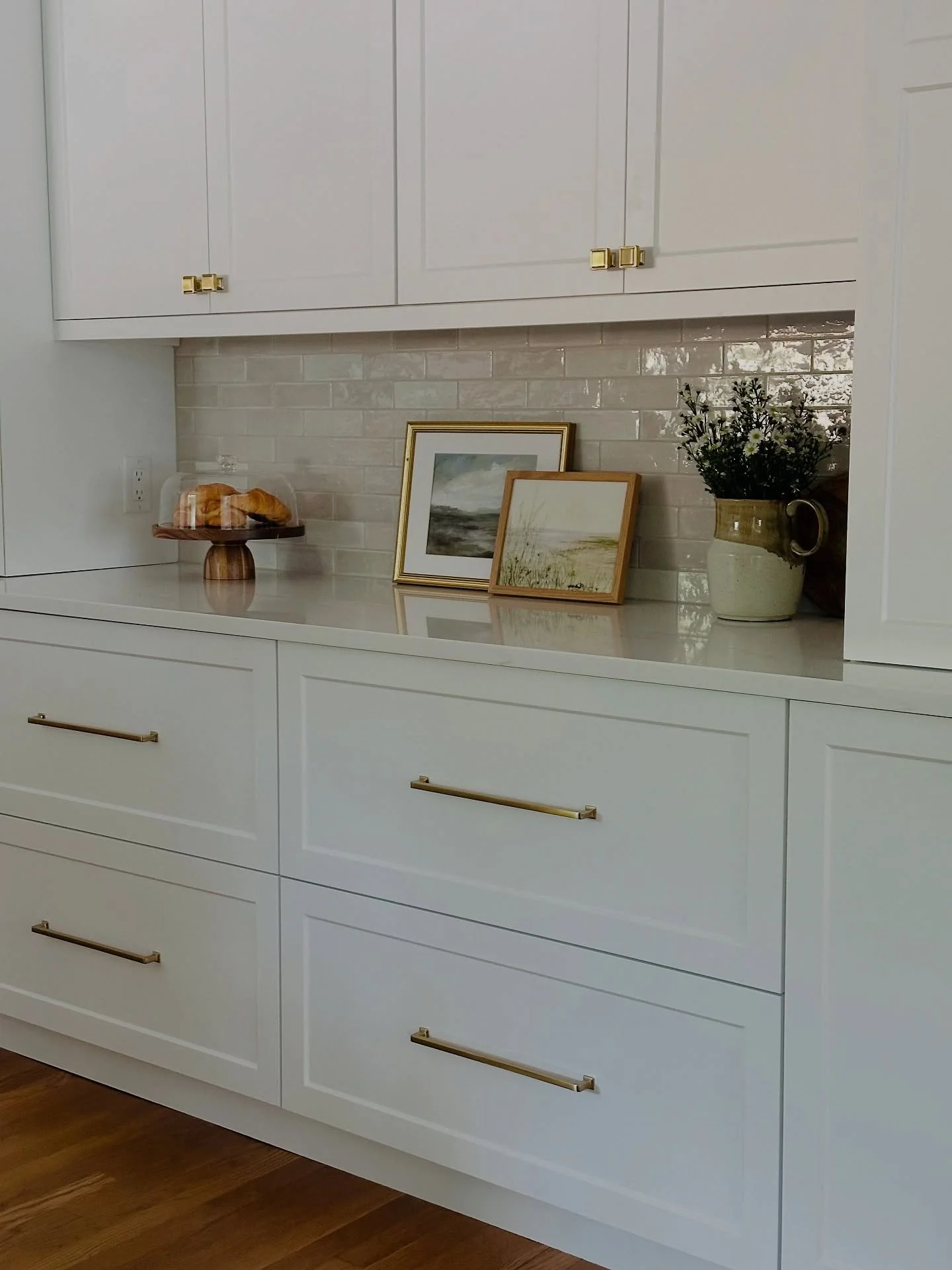 White built-in cabinet with four drawers and gold handles, a granite countertop, framed artwork, a vase with flowers, a cake stand with pastries, and a white subway tile backsplash. Heartwood Renovations. West Hartford, CT