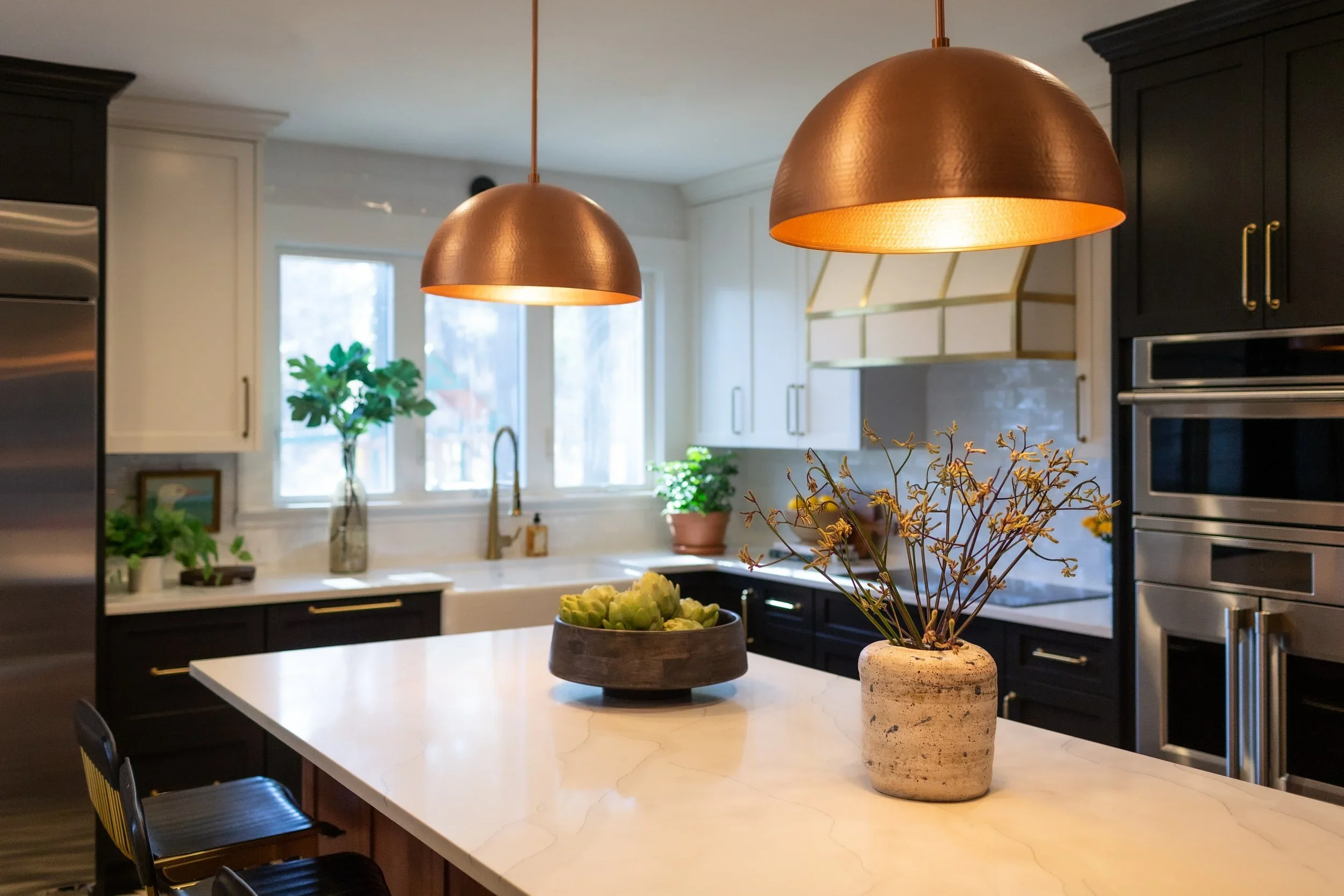 Modern kitchen with white and dark cabinets, stainless steel appliances, a large island with a white marble countertop, hanging copper pendant lights, and decorative plants and flowers. Heartwood Renovations. West Hartford, CT