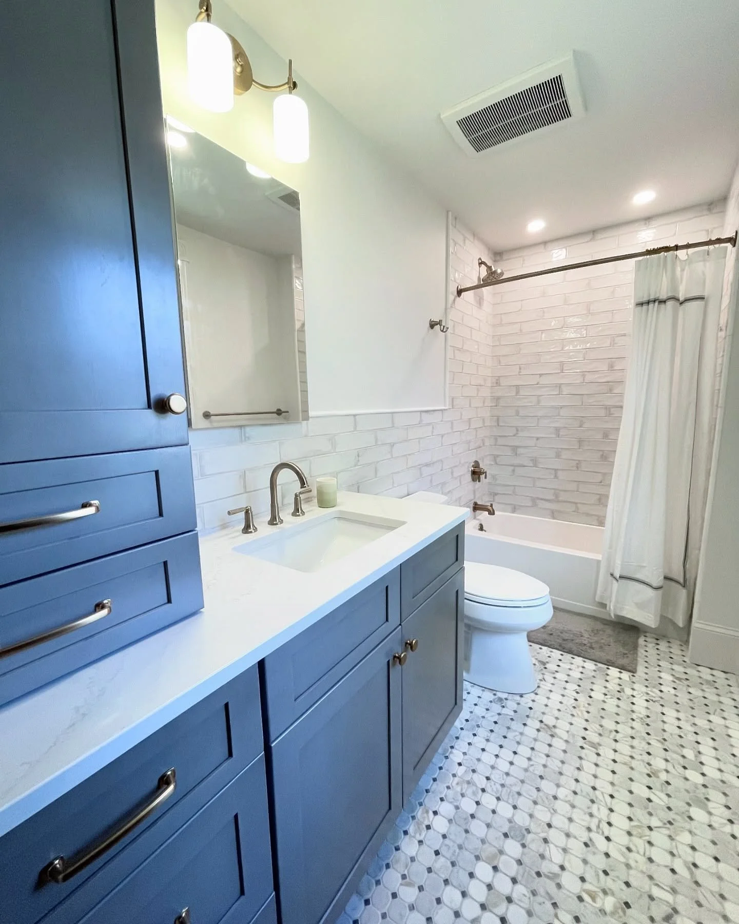 Modern bathroom with blue cabinetry, white countertop, mirror, wall-mounted light fixtures, white brick wall around the bathtub, shower curtain, toilet, and hexagonal tiled floor. Heartwood Renovations. West Hartford, CT