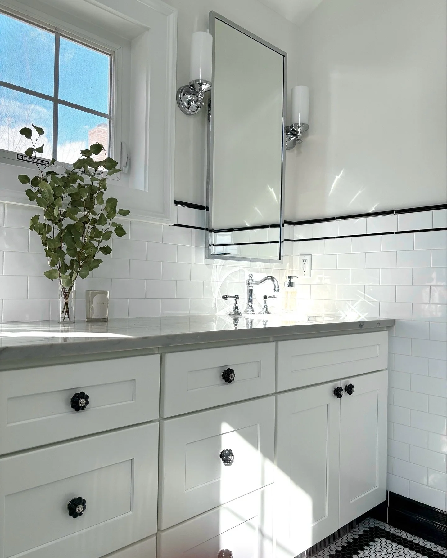 Bathroom with white cabinets, a marble countertop, a window, a mirror with a silver frame, wall-mounted light fixtures, a vase with greenery, and a hexagonal tile floor. Heartwood Renovations. West Hartford, CT