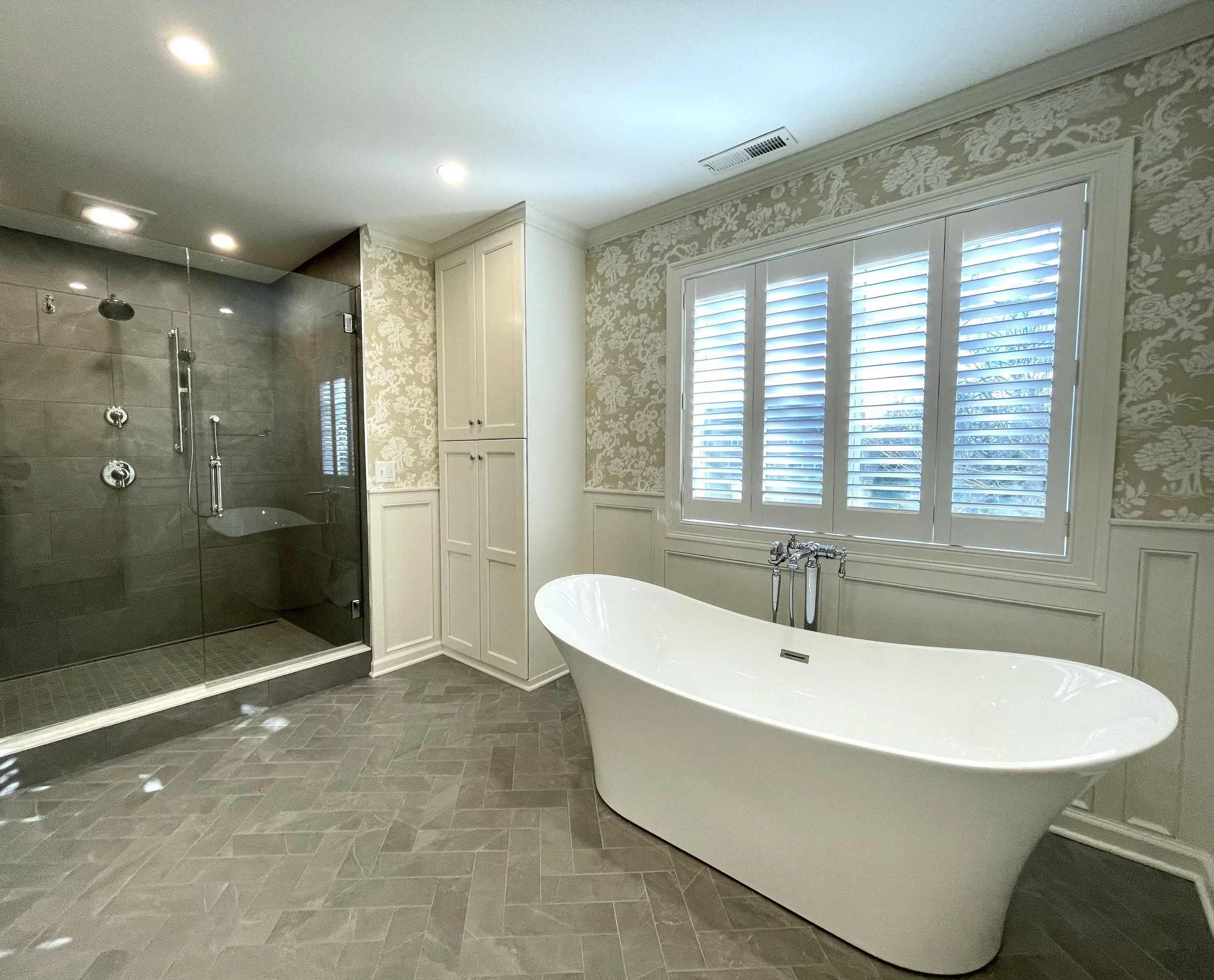 Modern bathroom with a freestanding bathtub, large window with shutters, and glass-enclosed shower with dark tiles. Heartwood Renovations. West Hartford, CT