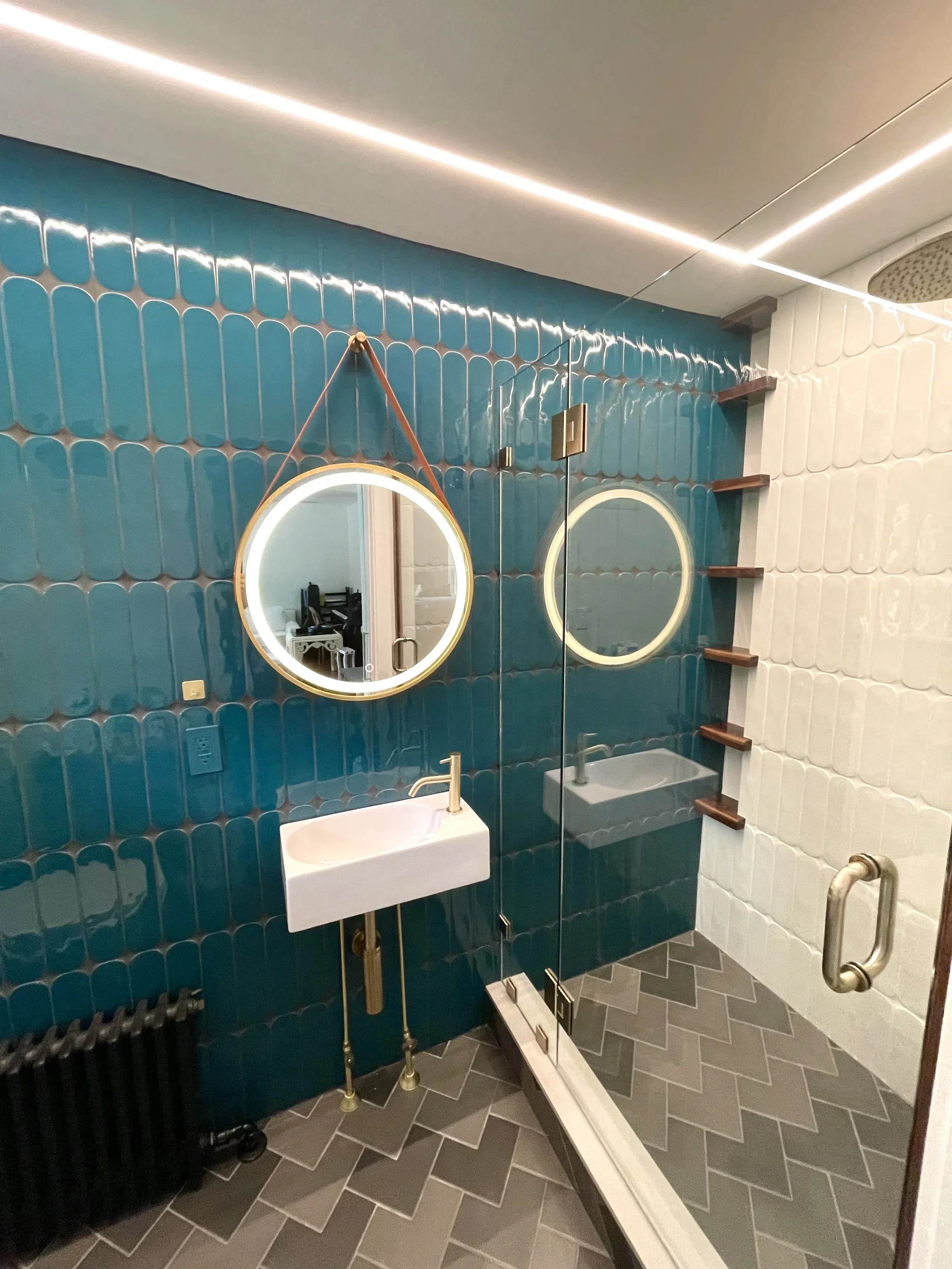 Modern bathroom with blue and white tiled walls, a small white sink with gold fixtures, round illuminated mirrors, glass shower enclosure with a brass handle, and wood corner shelves. The floor has a herringbone pattern. Heartwood Renovations. West H