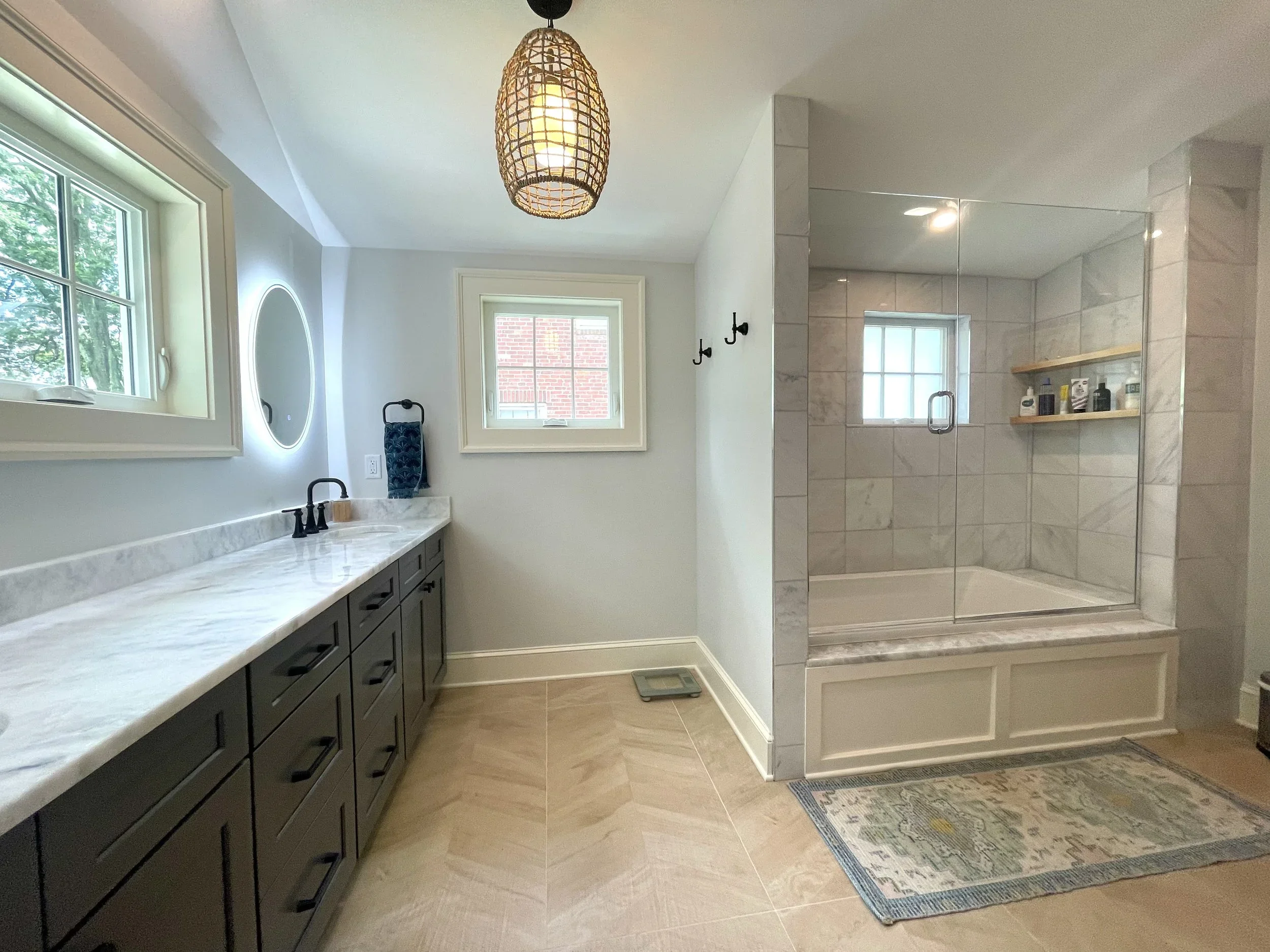 Bathroom with light grey walls, marble countertop vanity with black fixtures, two small windows, round mirror with backlight, beige tile flooring, and a large walk-in shower with glass doors, marble tiles, a built-in shelf, and small window. A rug and wall hooks complete the space.