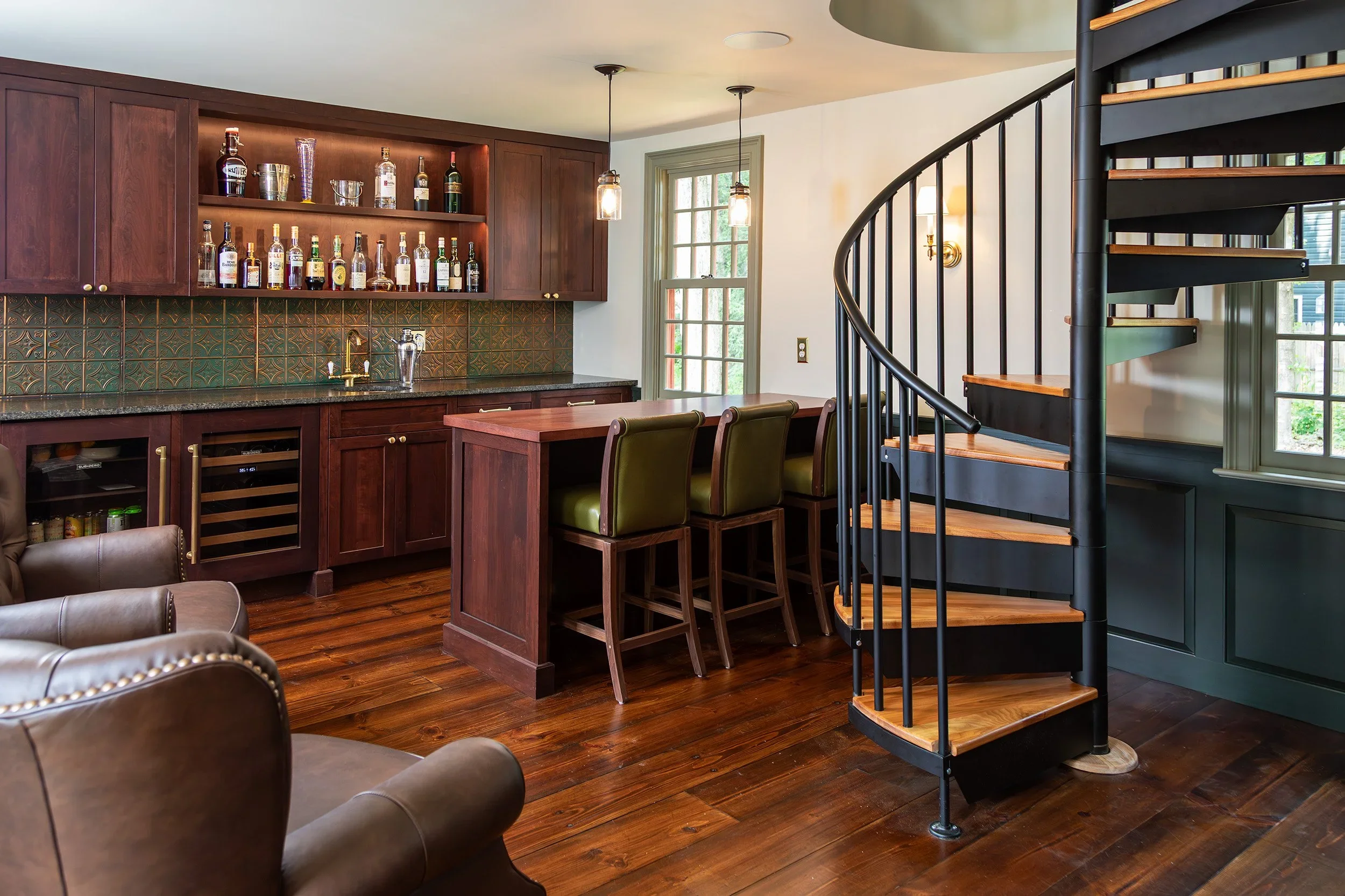 Home bar area with dark wood cabinets, a built-in wine fridge, and a countertop with liquor bottles. There are three green bar stools at a wooden counter, hardwood floors, and a black spiral staircase with wooden steps. Natural light enters through a window and a glass door.