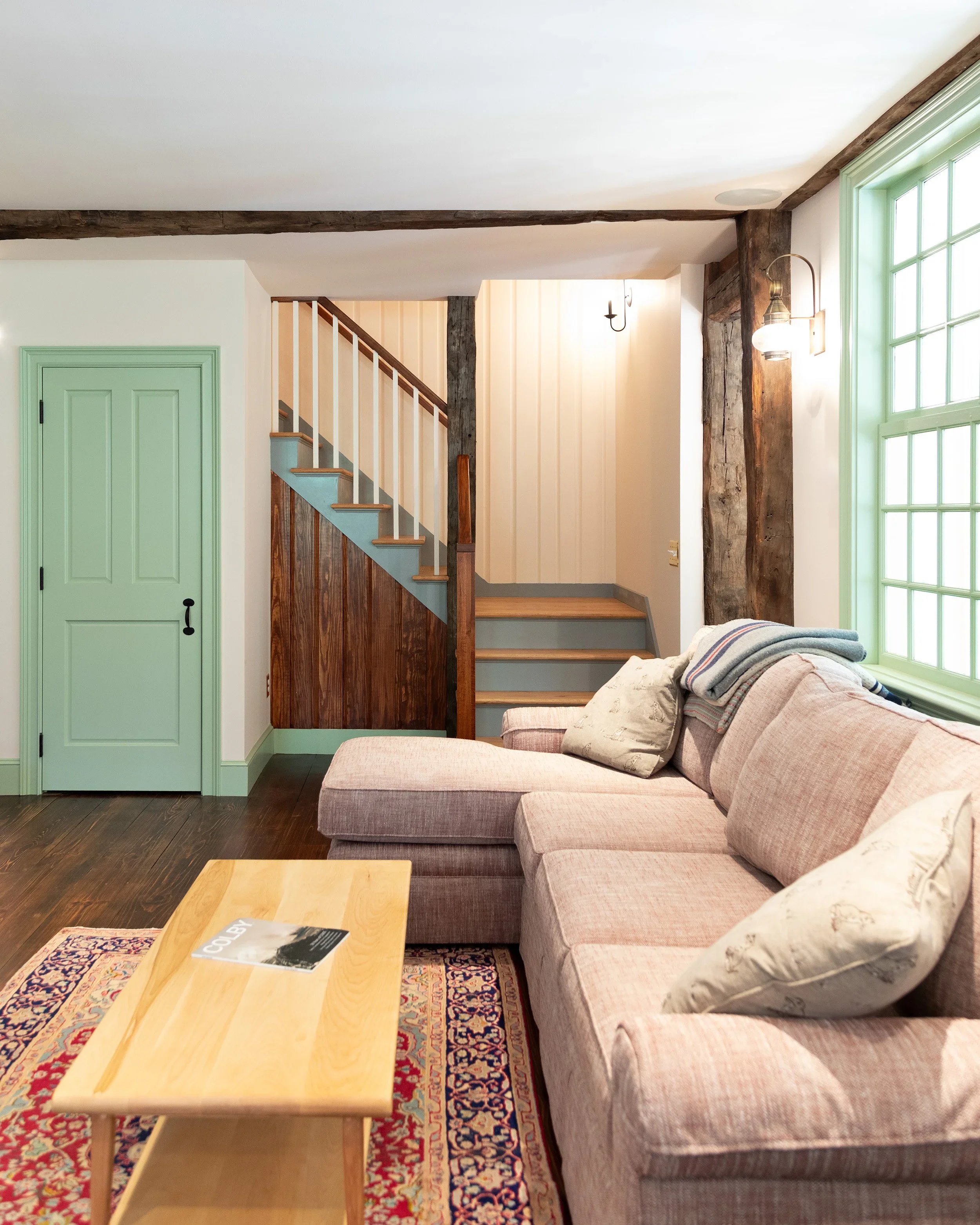 Cozy living room with pink sectional sofa, throw pillows, wooden coffee table, large window with green trim, stone and wood wall accents, staircase, and soft lighting. Heartwood Renovations. West Hartford, CT