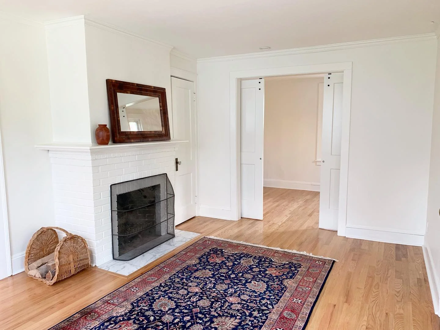 Living room with white painted brick fireplace, gold and blue patterned rug, and a wicker basket near the hearth. There is a mirror and a small clay pot on the mantle. The room has hardwood floors and white walls with crown molding. An open doorway with white trim leads to another room.