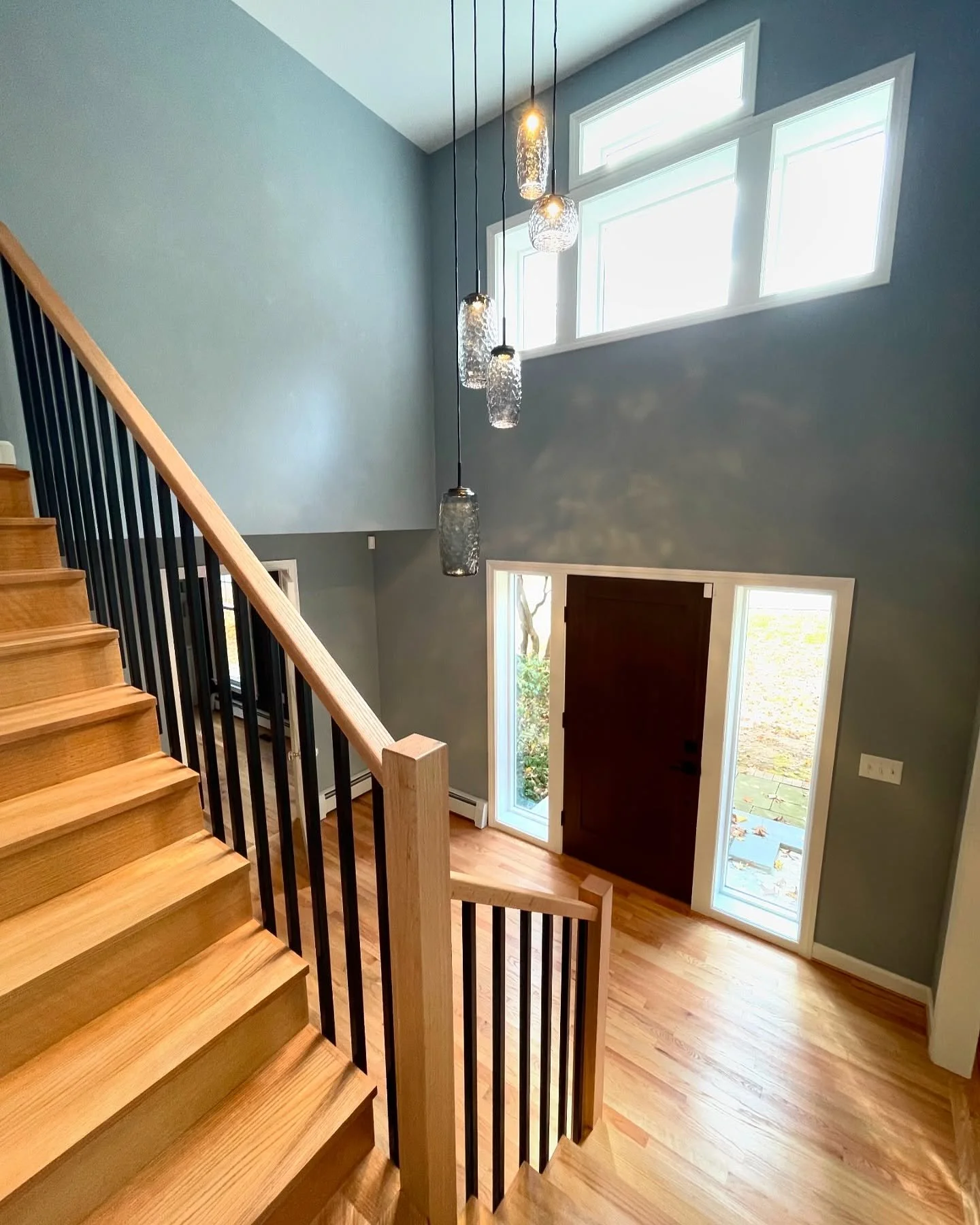 Interior view of a modern home entrance with wooden stairs, hanging pendant lights, large windows, and a dark front door. Heartwood Renovations. West Hartford, CT