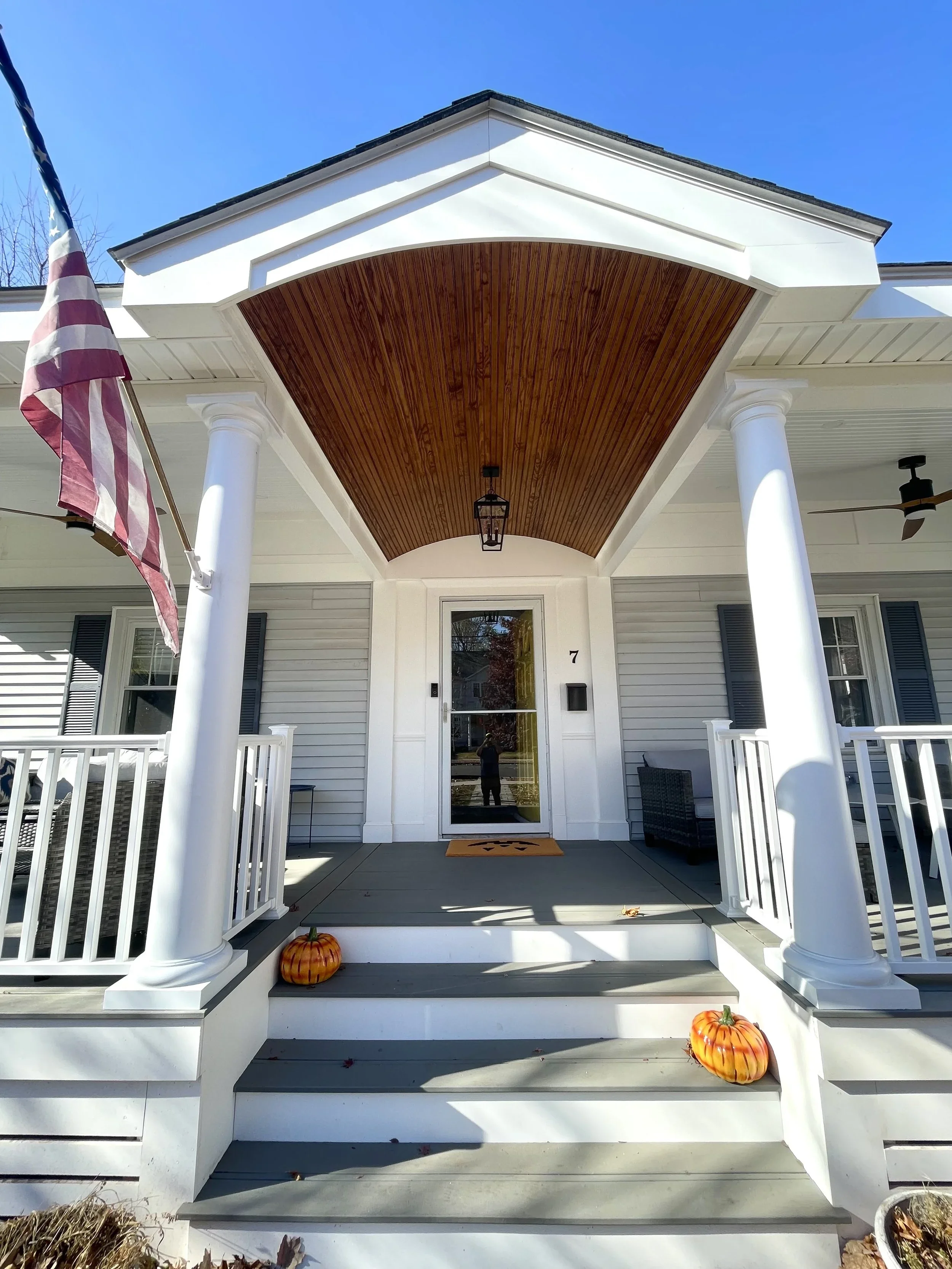 Front porch of a house with white railings and columns, two orange pumpkins on the steps, and an American flag on the left side. The house has a gray exterior, white trim, and a wooden ceiling on the porch. Heartwood Renovations. West Hartford, CT