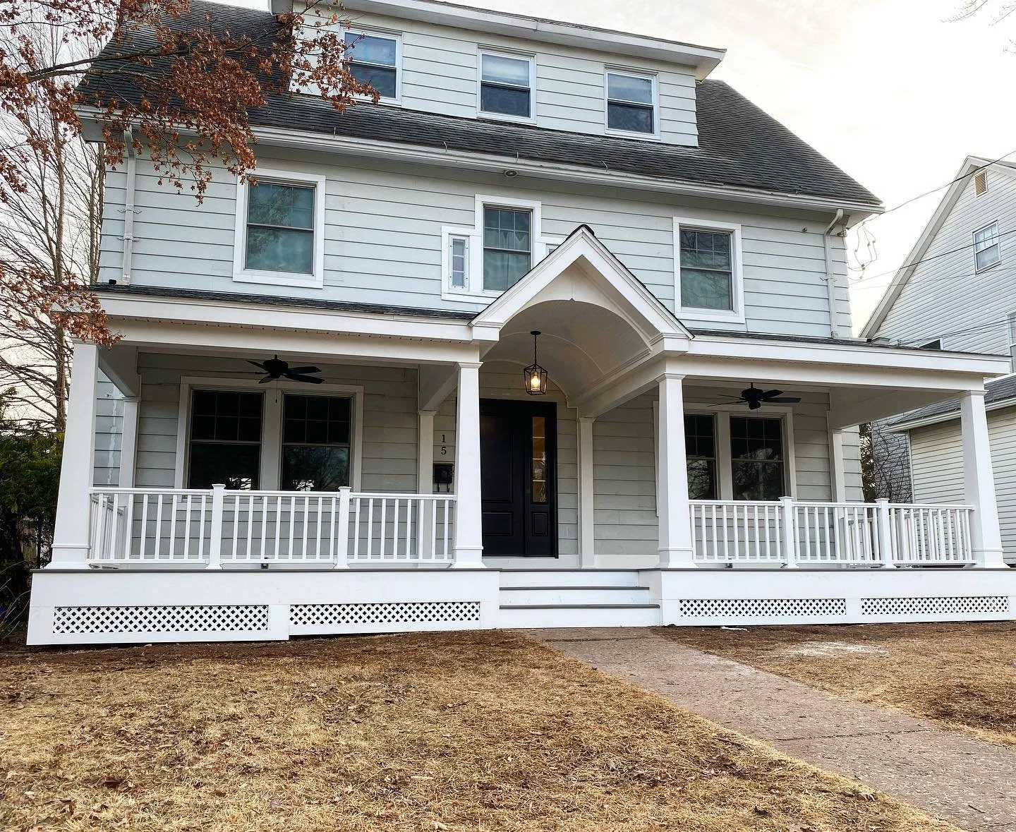 A white three-story house with a front porch, black front door, and a gable roof. The porch has white railings, two ceiling fans, and a hanging lantern. The house has multiple windows, some with white shutters. There is a small concrete walkway leading to the front steps, and the yard has brownish grass.