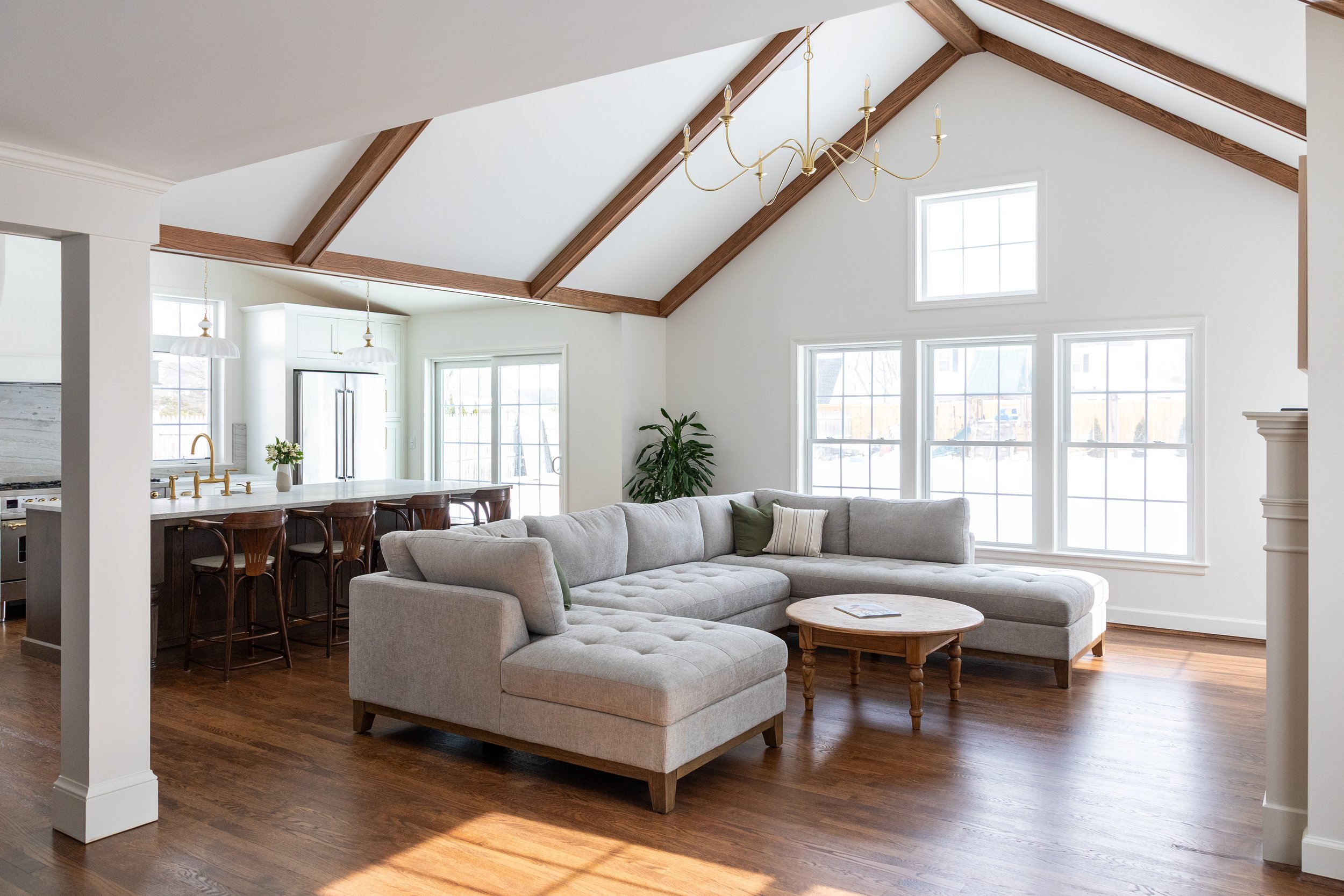 Bright living room with white walls, vaulted ceiling with wooden beams, large windows, a beige sectional sofa, wooden coffee table, hardwood floors, and a chandelier. Heartwood Renovations. West Hartford, CT