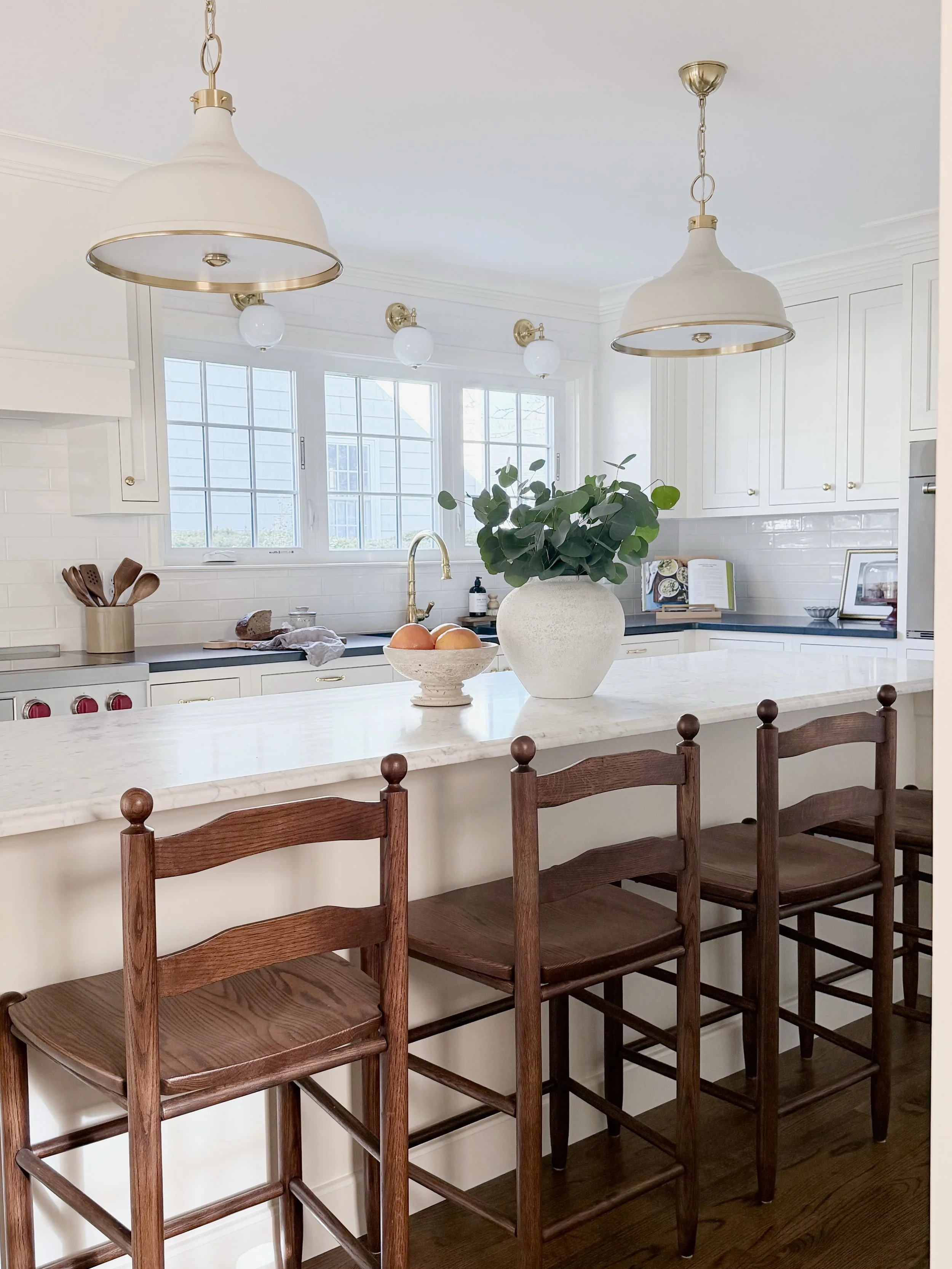 Bright white kitchen with white cabinets, marble countertop island, large window, green plant in white vase, wooden bar stools, and gold and white pendant lights. Heartwood Renovations. West Hartford, CT