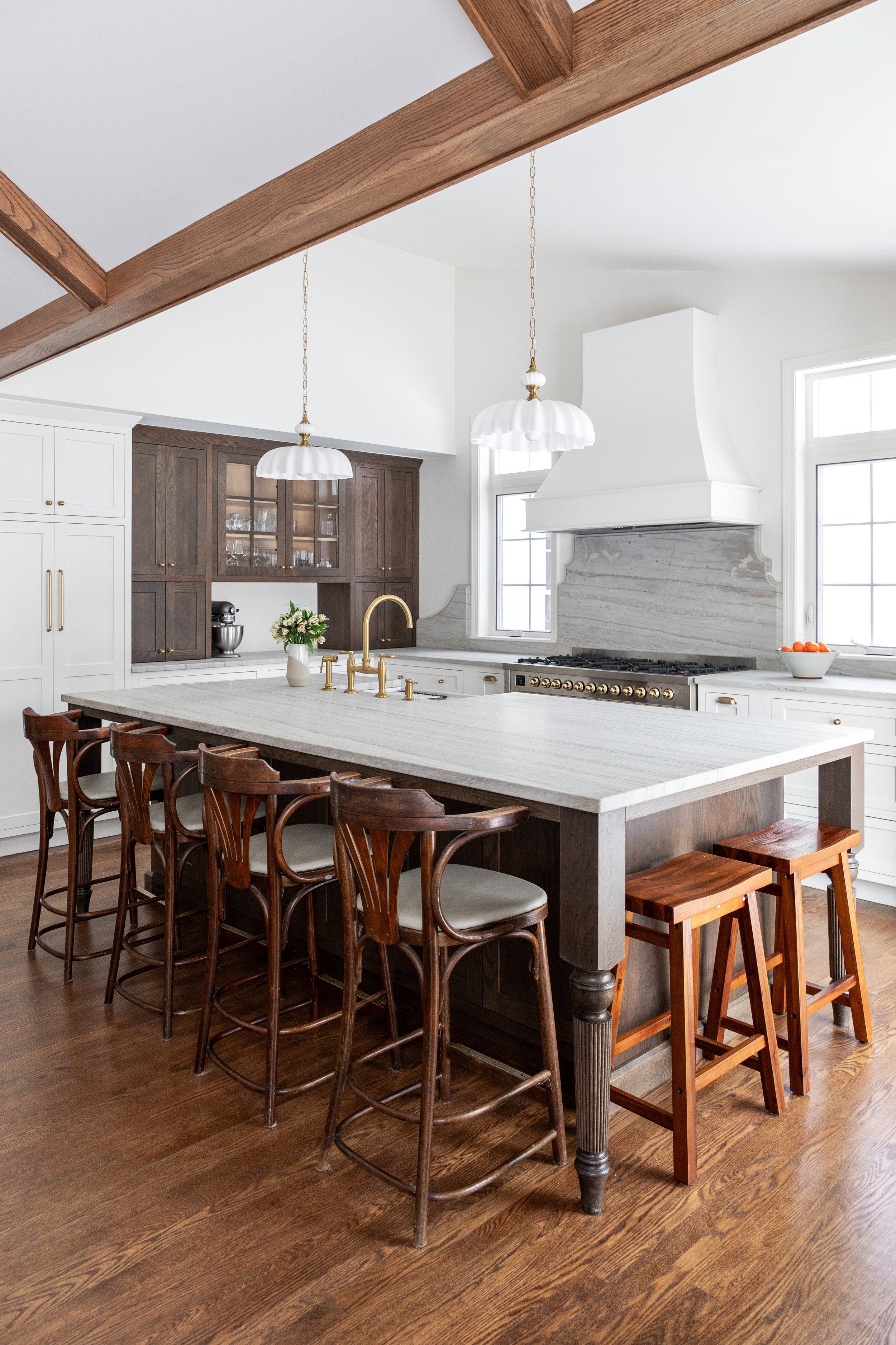 Modern kitchen with a large white island, wooden stools, white and dark wood cabinetry, a range hood, pendant lights, and hardwood floors. Heartwood Renovations. West Hartford, CT
